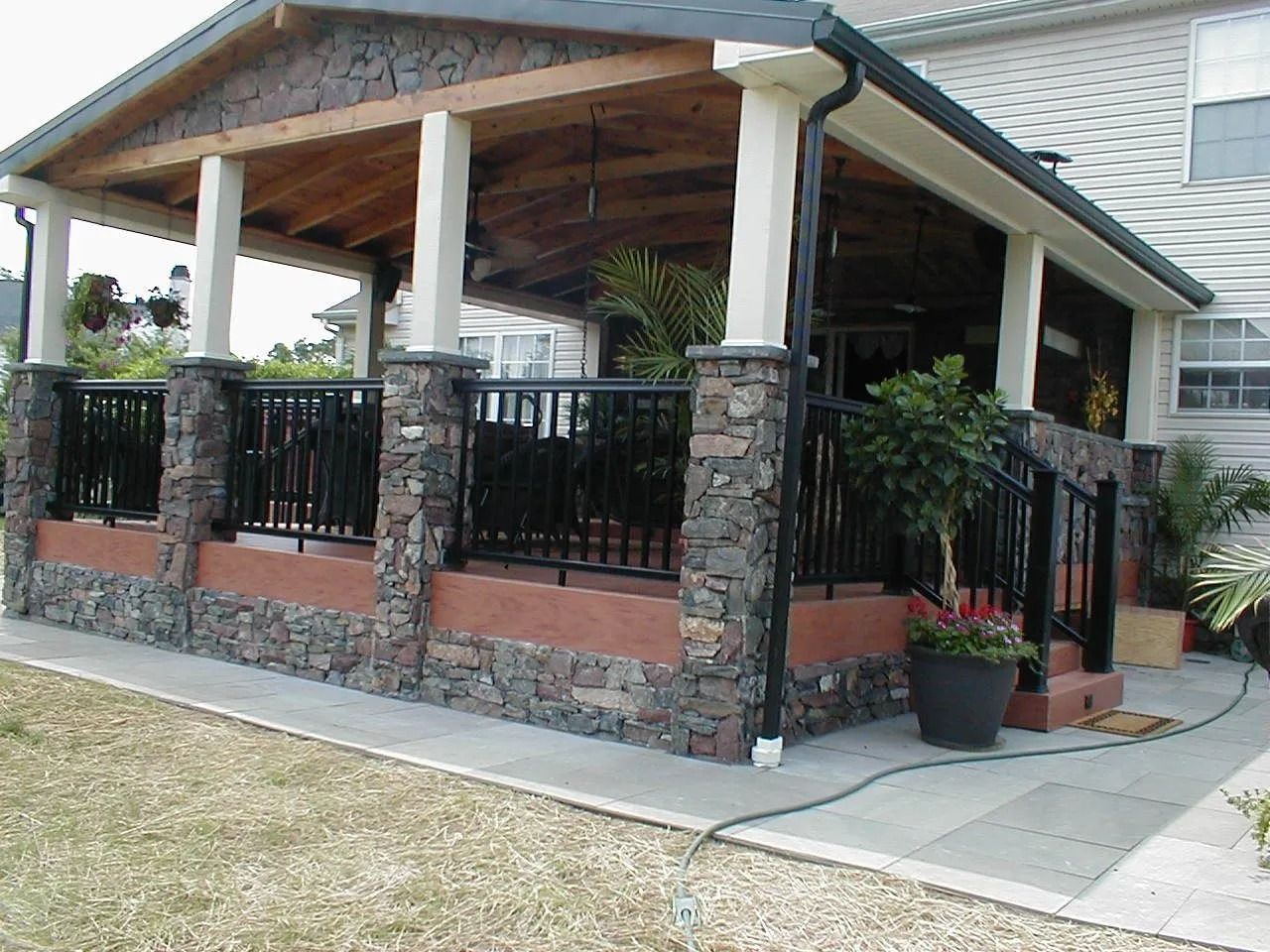 Covered porch with stone columns and black railing, adjacent to a house.