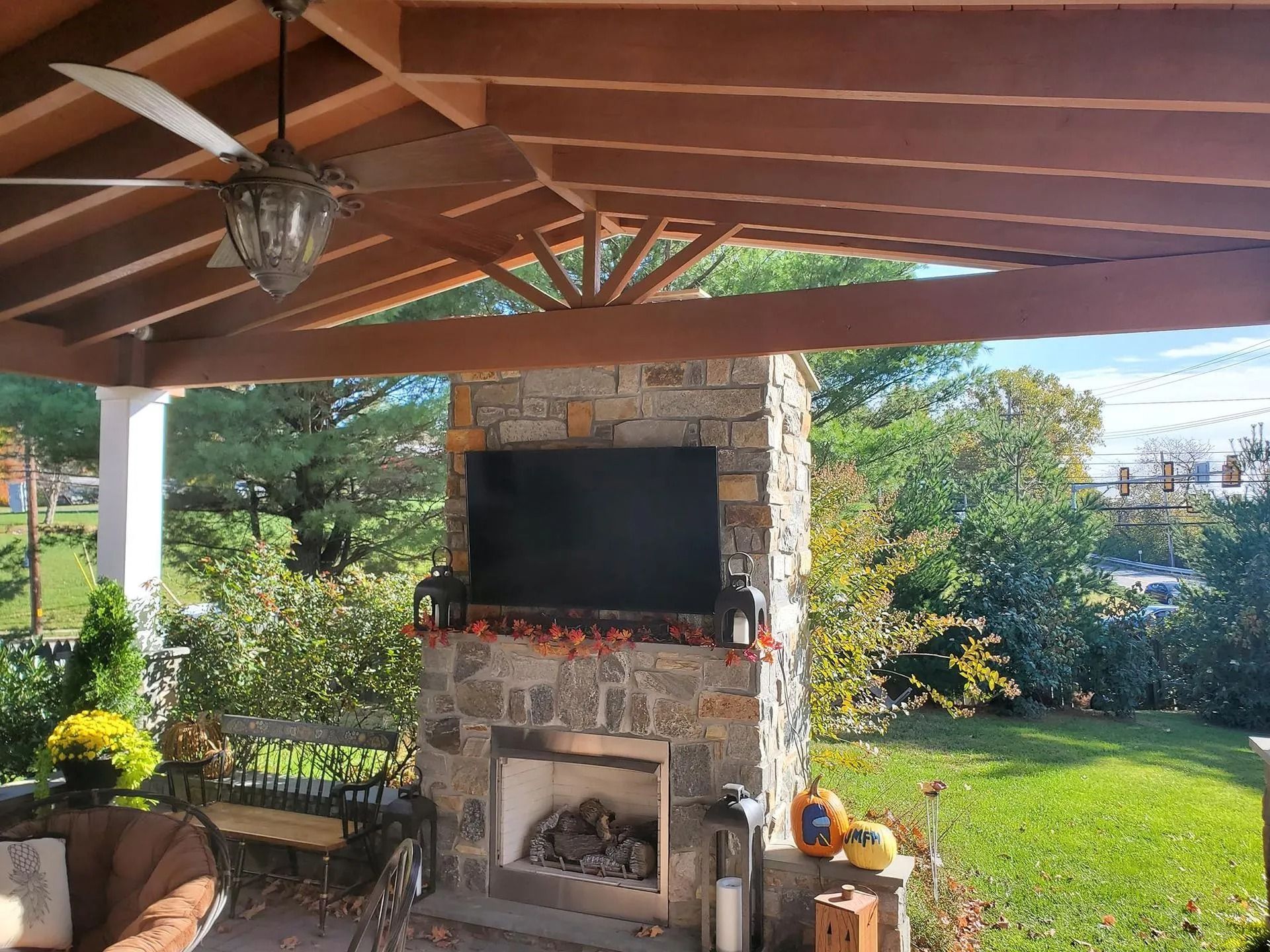 Outdoor fireplace with a mounted TV under a covered patio, surrounded by foliage and a ceiling fan.