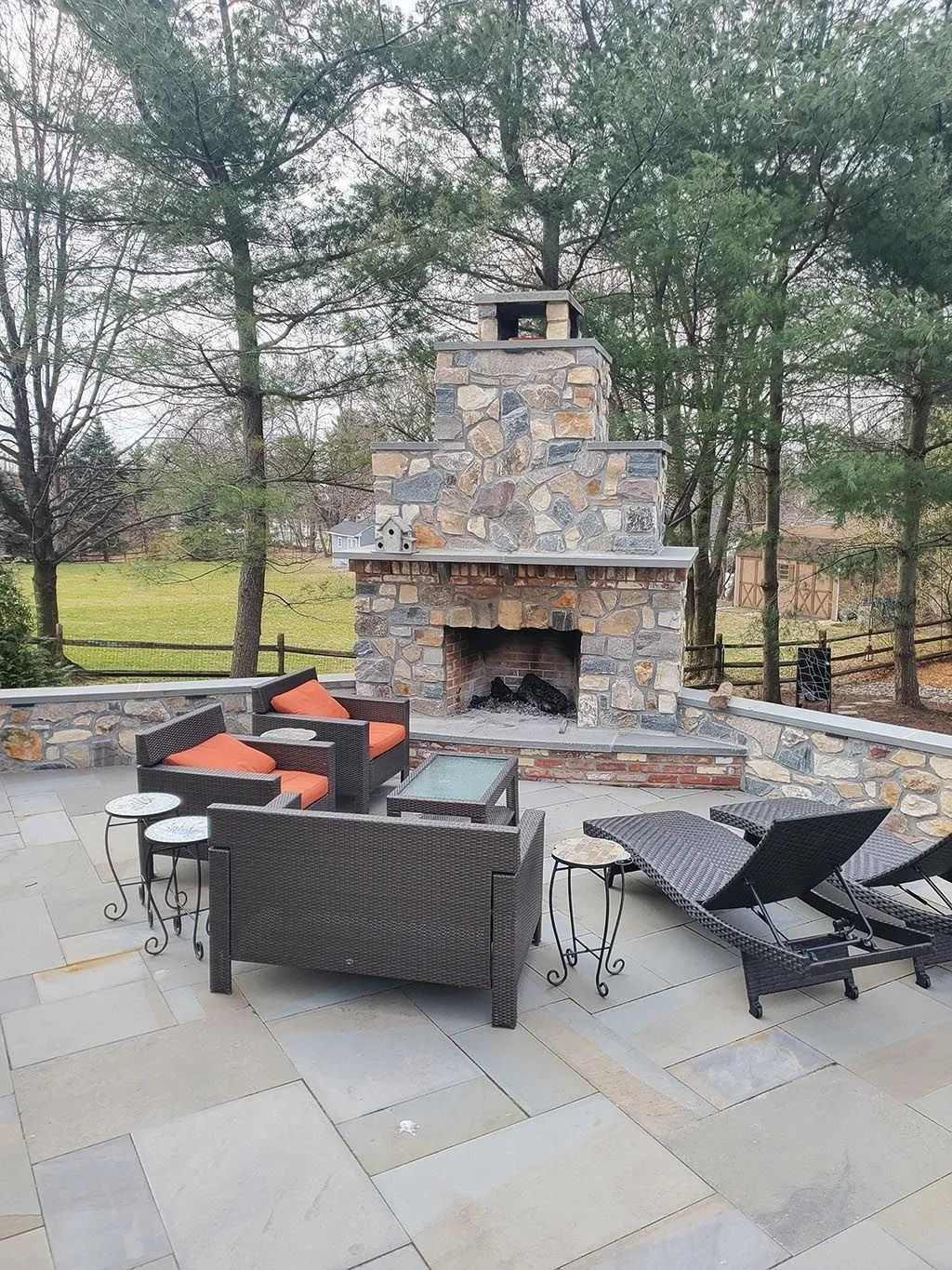 Outdoor stone fireplace with seating area; patio with lounge chairs. Trees in the background.