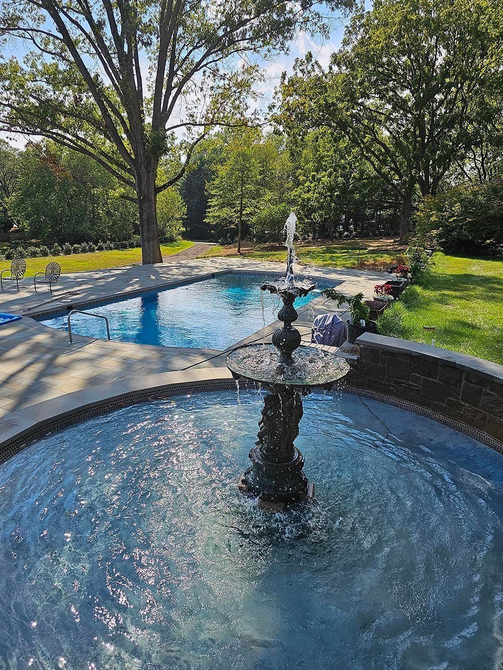 Fountain sprays water into a pool, with a larger pool and trees in the background. Sunny day.