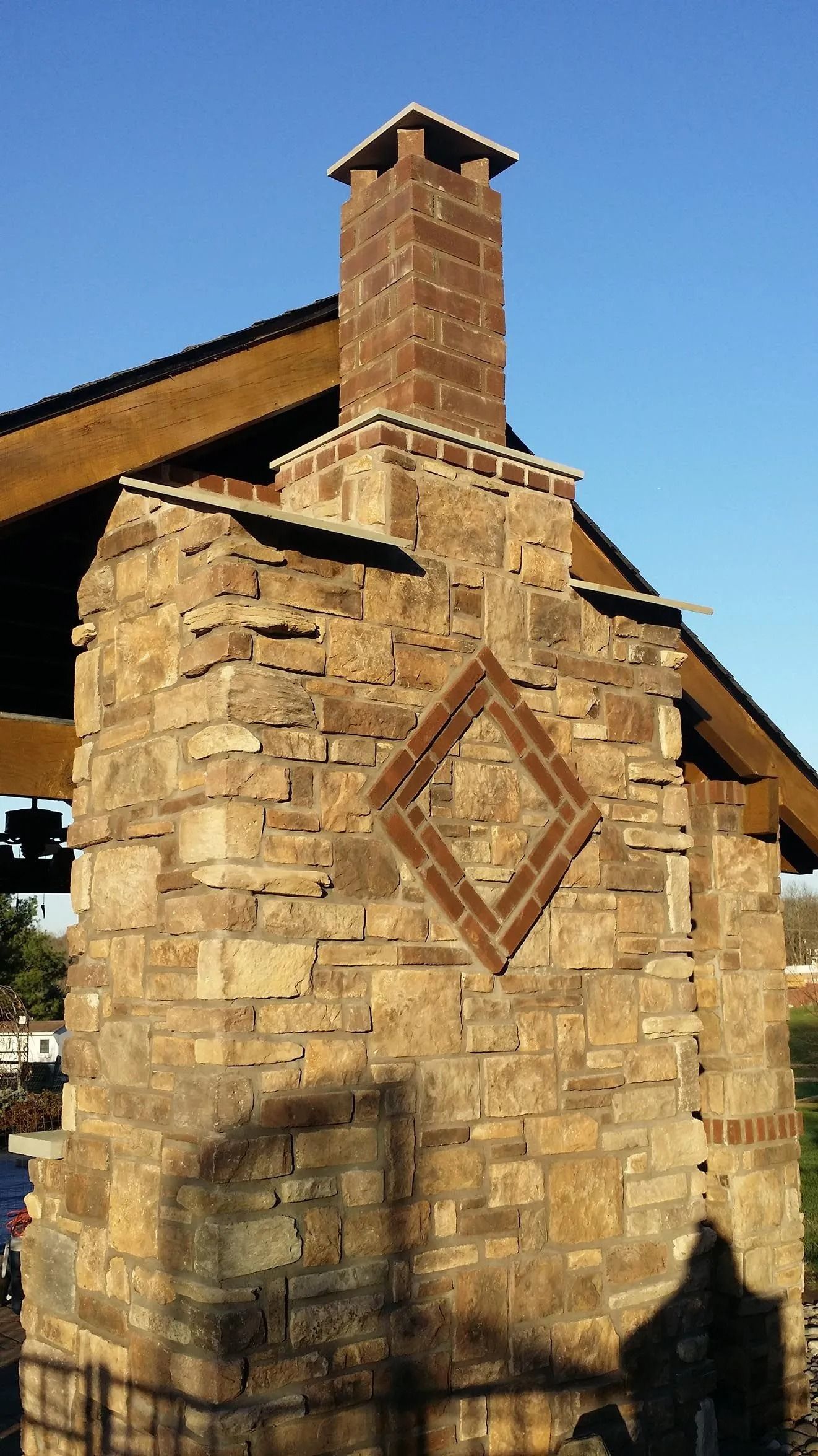 Stone chimney with a diamond pattern, attached to a brown wooden roof structure against a blue sky.