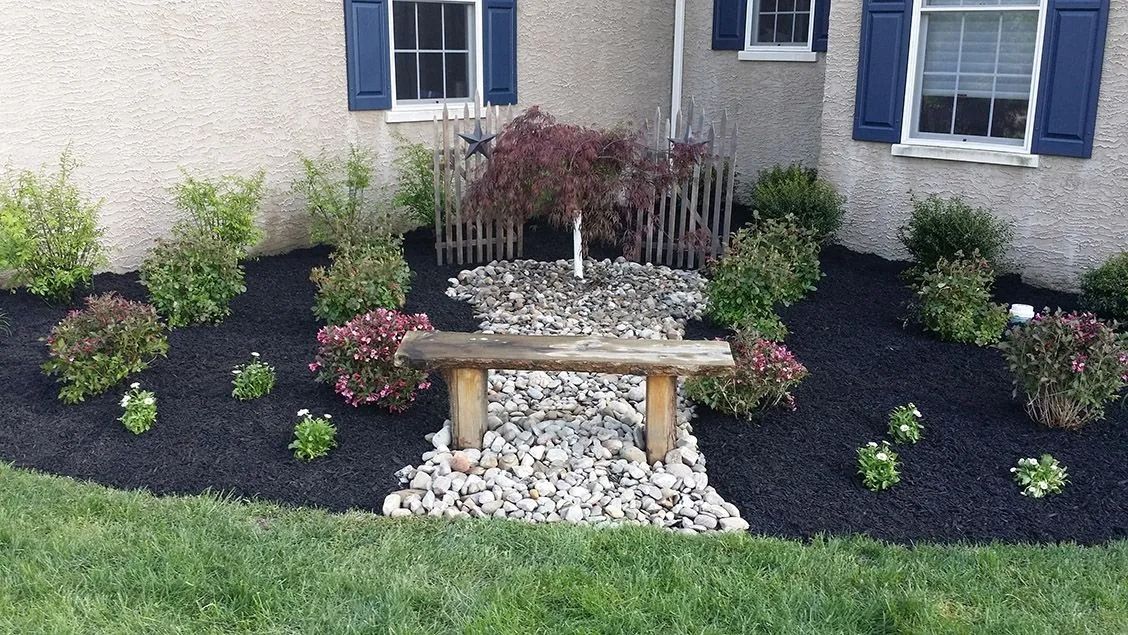 Front yard with a rock garden, bench, and various shrubs and plants on dark mulch.