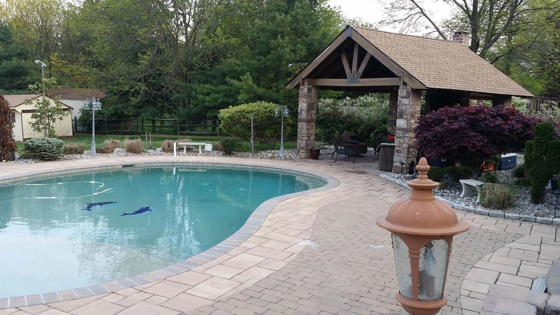 Pool with stone patio, gazebo, and surrounding trees.