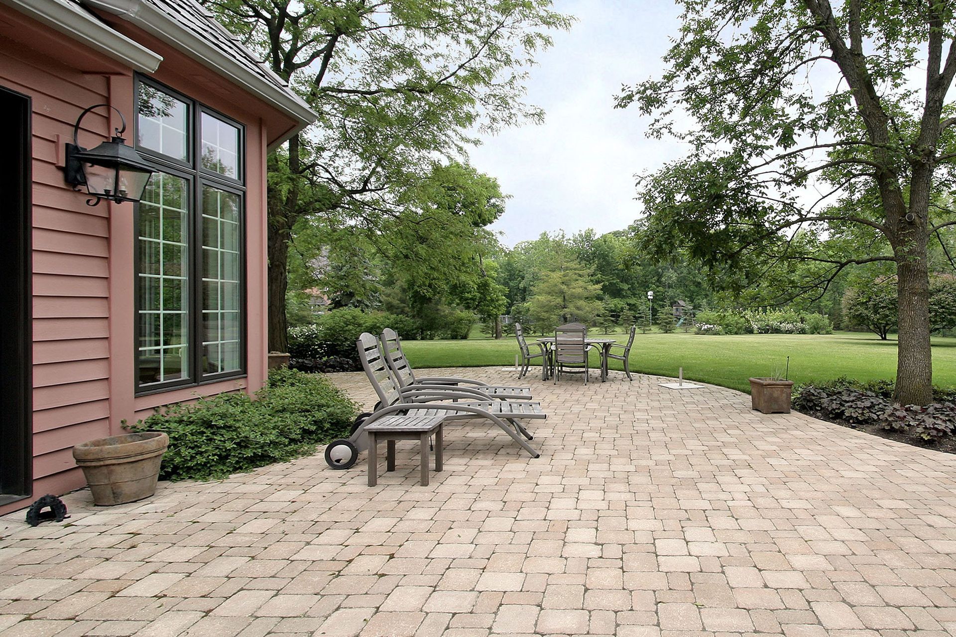 Brick patio with lounge chairs near a house with large windows, overlooking a green lawn with trees.