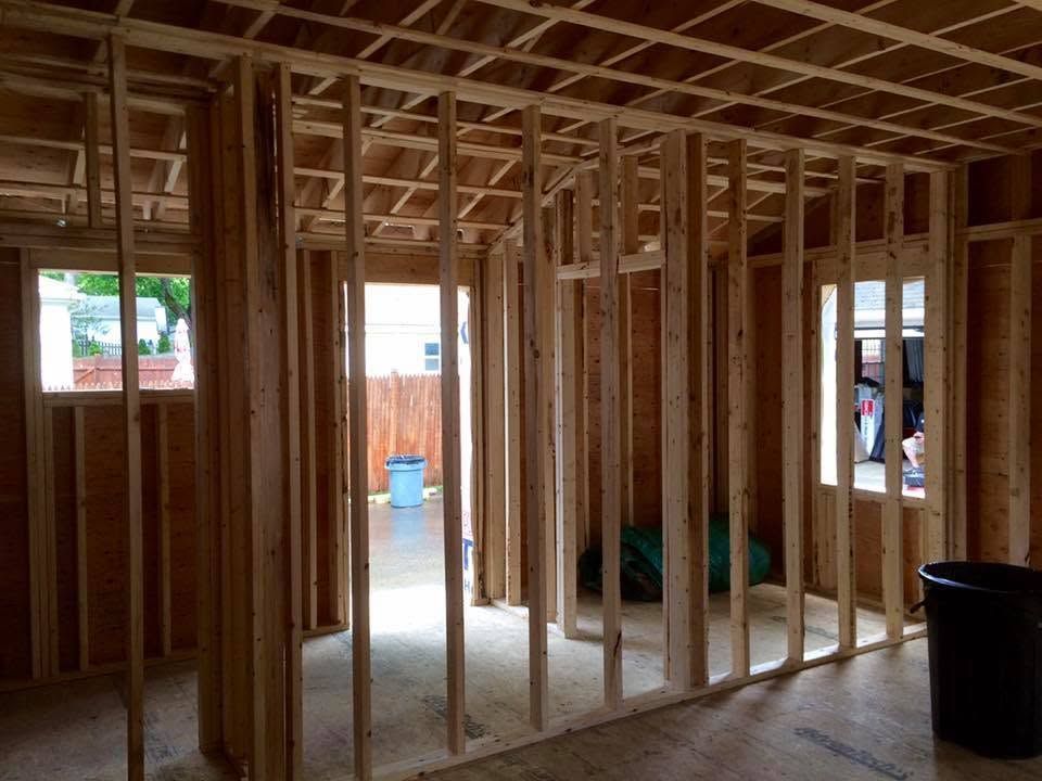 Unfinished house interior with exposed wooden framing, open window, and black trash bin on the right.