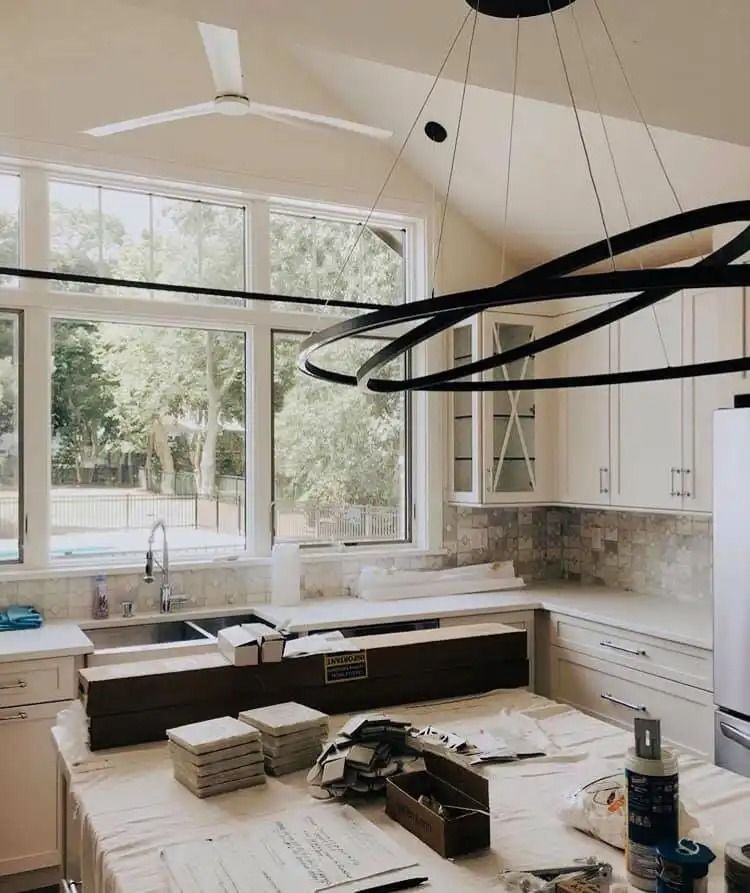 Bright modern kitchen under renovation with white cabinets, large windows, and materials on the unfinished island.