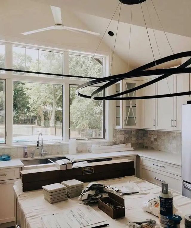 Bright unfinished kitchen with large windows, white cabinets, and materials spread across the island.