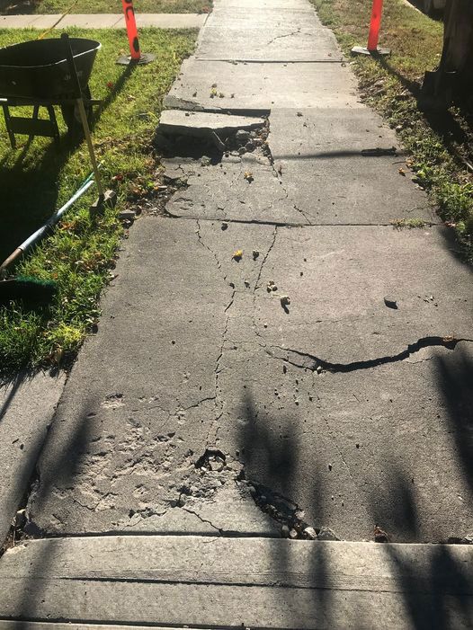A concrete sidewalk with a hole in it and a wheelbarrow in the grass.