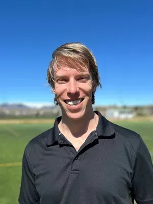 Smiling person in a black polo shirt standing outdoors on a sunny field with blue sky.