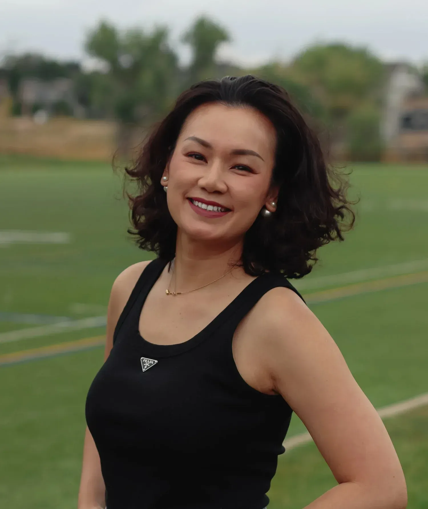 Smiling woman in a black tank top standing on a grassy field outdoors.