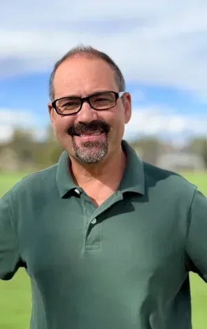 Smiling man in glasses and green polo shirt standing outdoors with a blurred sky and field behind him