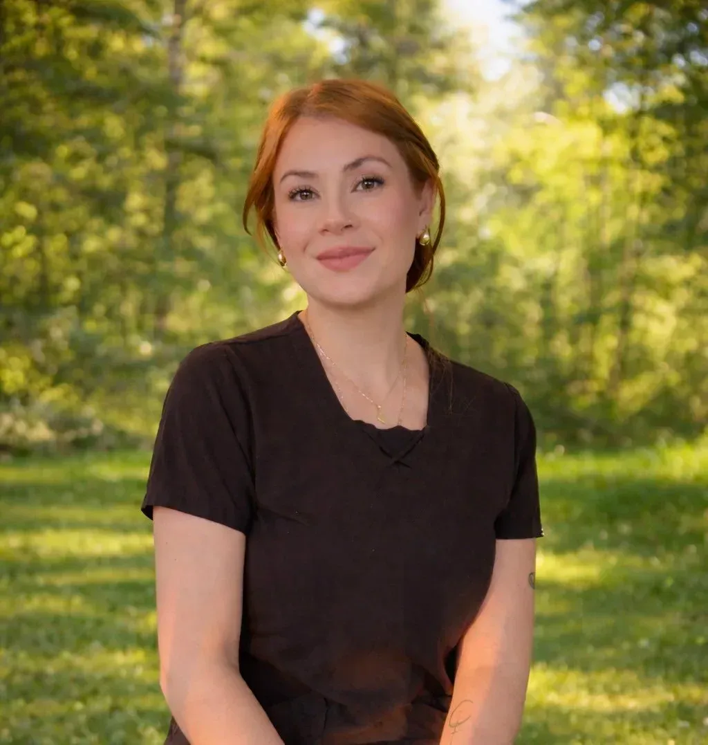 A woman in a black shirt sits outdoors in a sunlit park with green trees behind her.