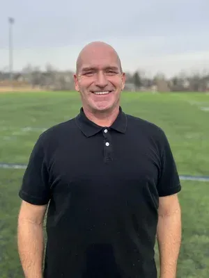 Smiling man in a black polo standing on a green field with blurred trees in the background
