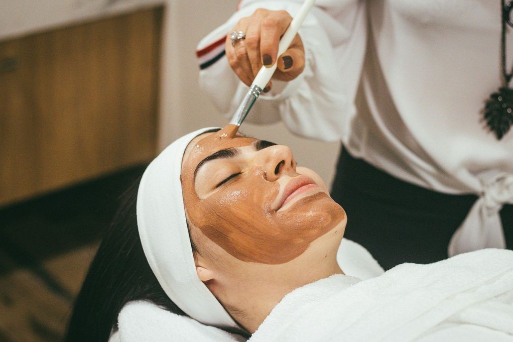 Esthetician applying facial treatment with a brush to a client lying on a spa bed.