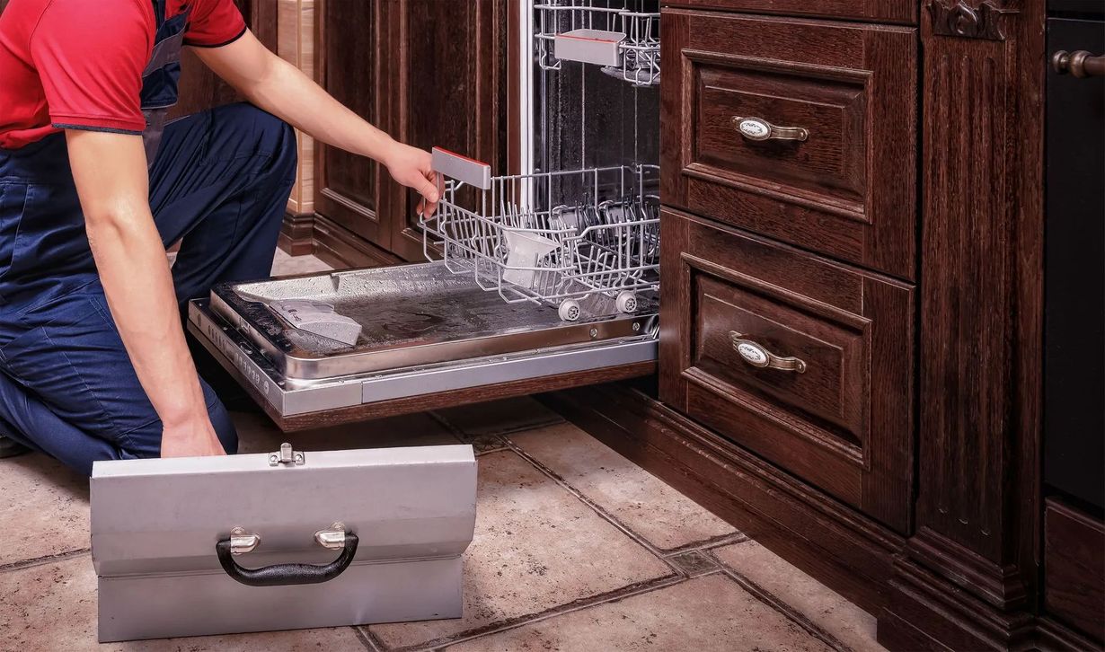 Person in blue overalls repairs a dishwasher in a dark wood kitchen; toolbox on the floor.