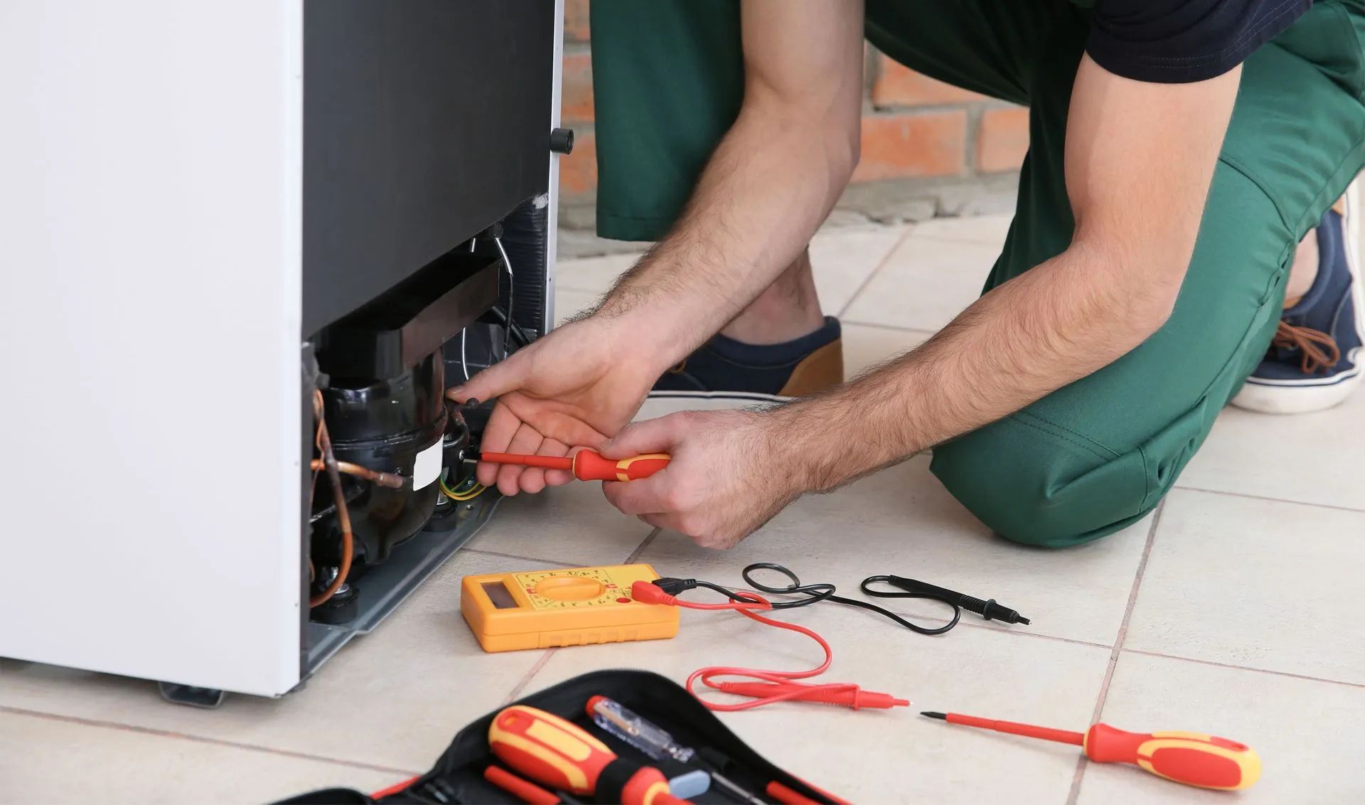 A repairman in green overalls tests a refrigerator with a multimeter and screwdriver on a tile floor.