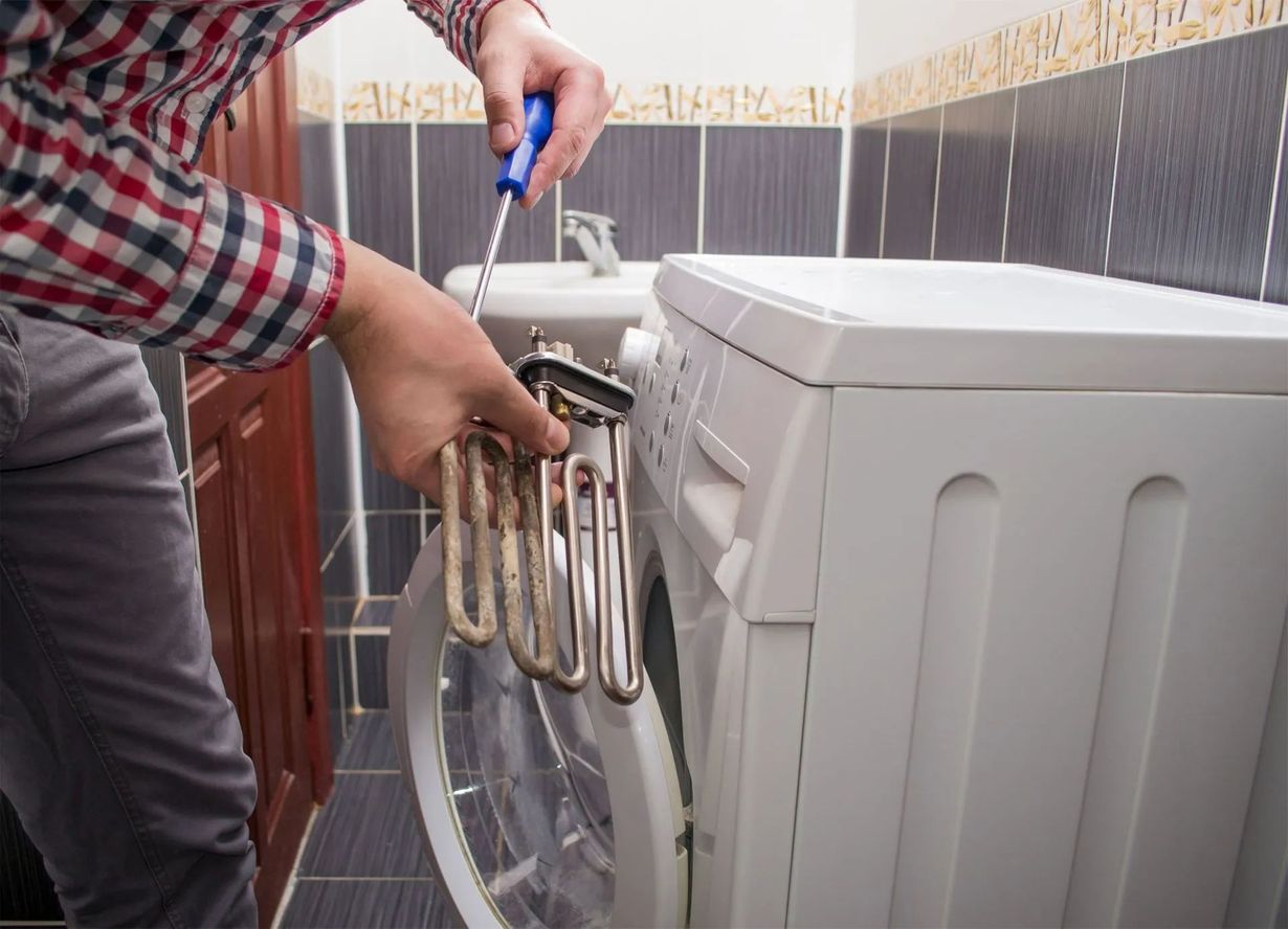 Man repairs washing machine in bathroom, holding a heating element with a screwdriver.