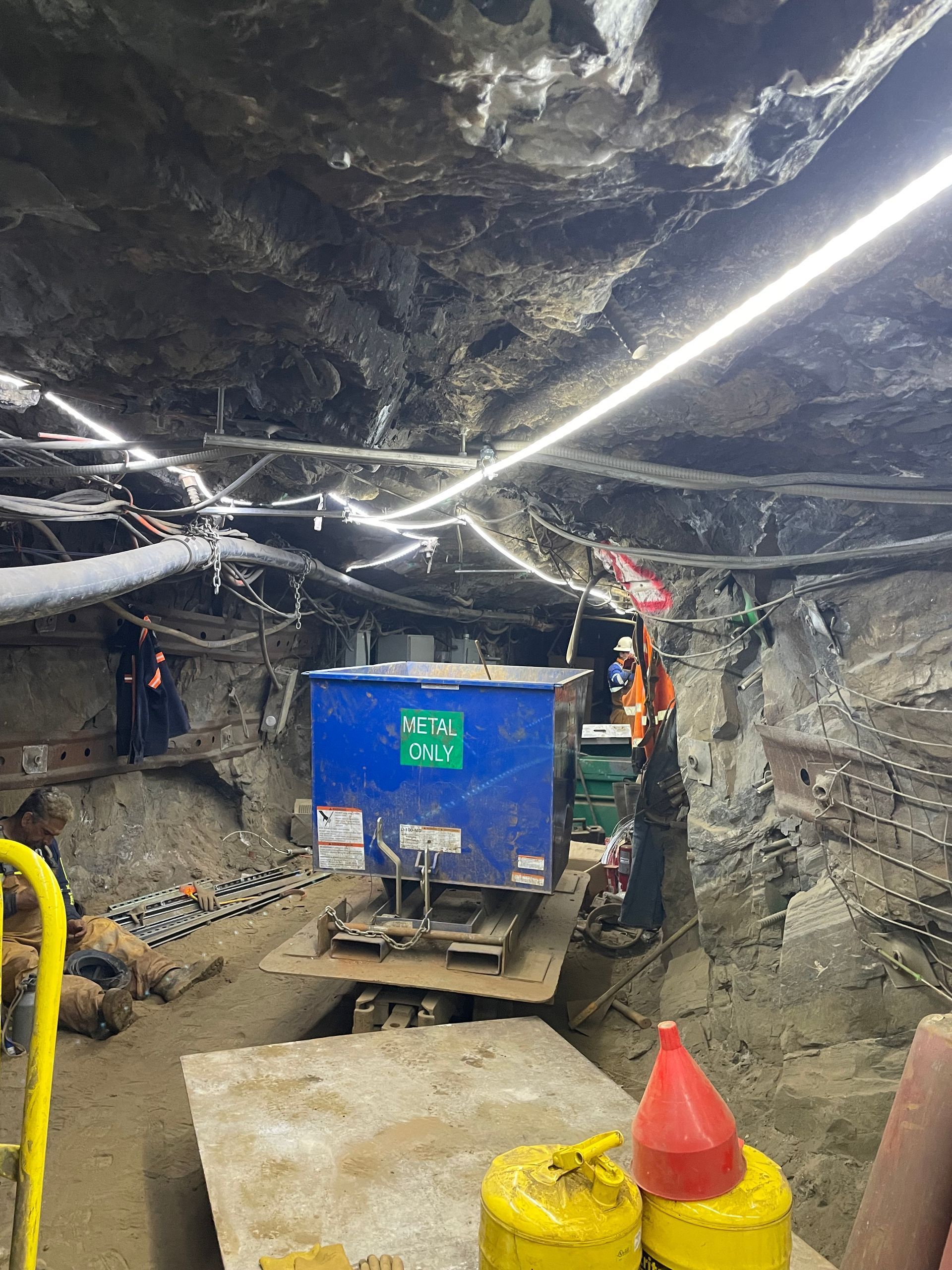 Construction tunnel with blue equipment on a platform; workers near the machinery, lights illuminate the work area.