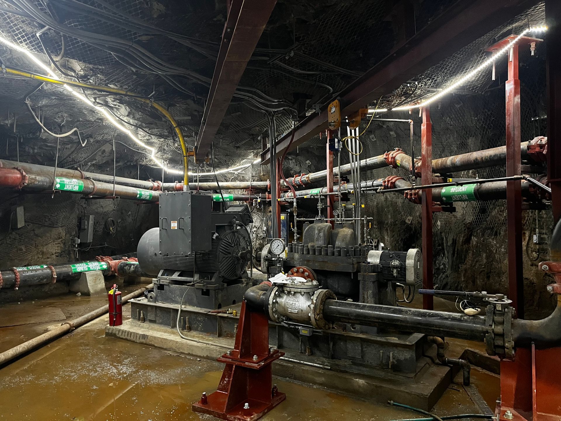 Machinery and pipes in an underground tunnel, illuminated by overhead lights.
