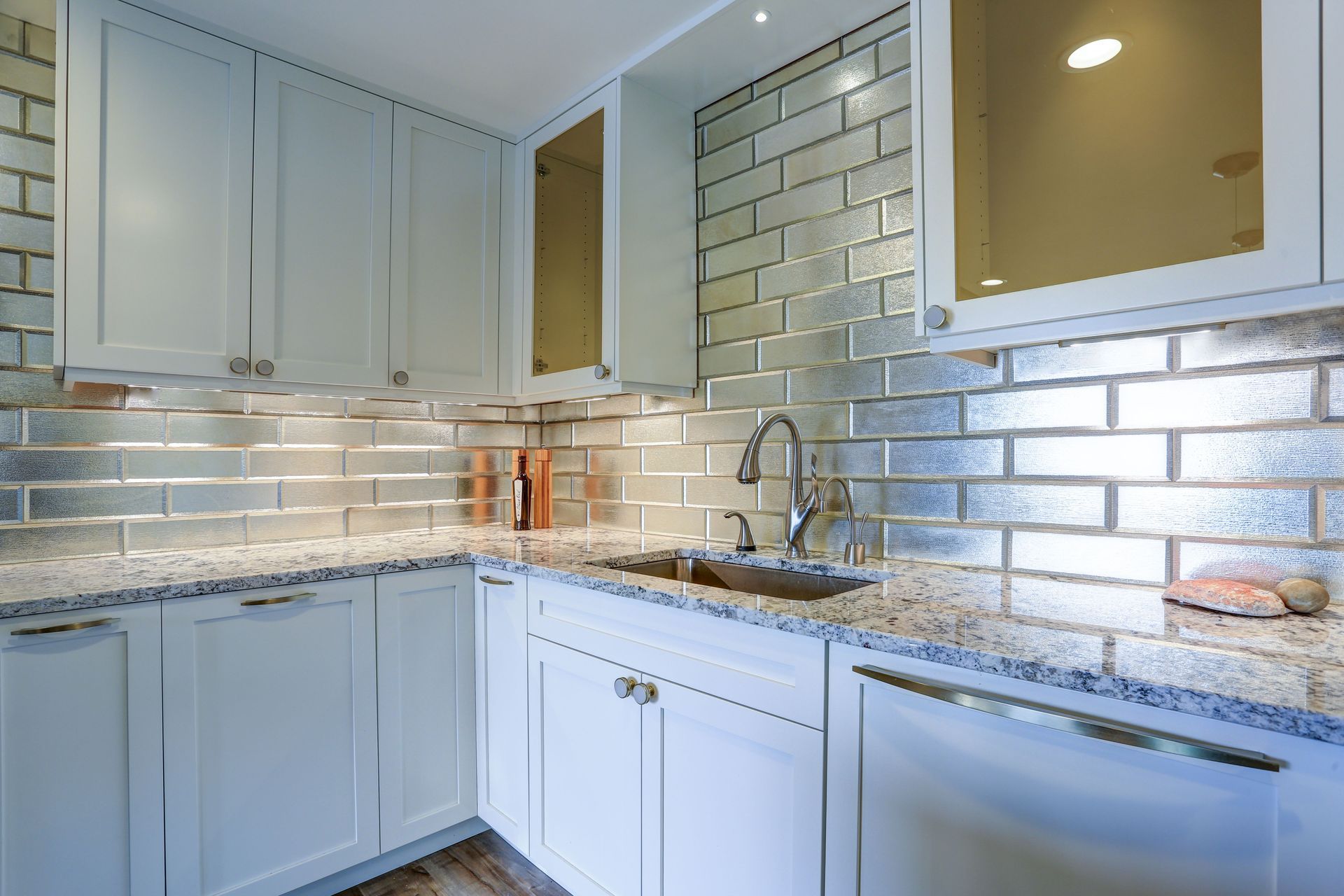 A bright kitchen featuring white cabinets, granite countertops, and a reflective, metallic-tiled subway backsplash.
