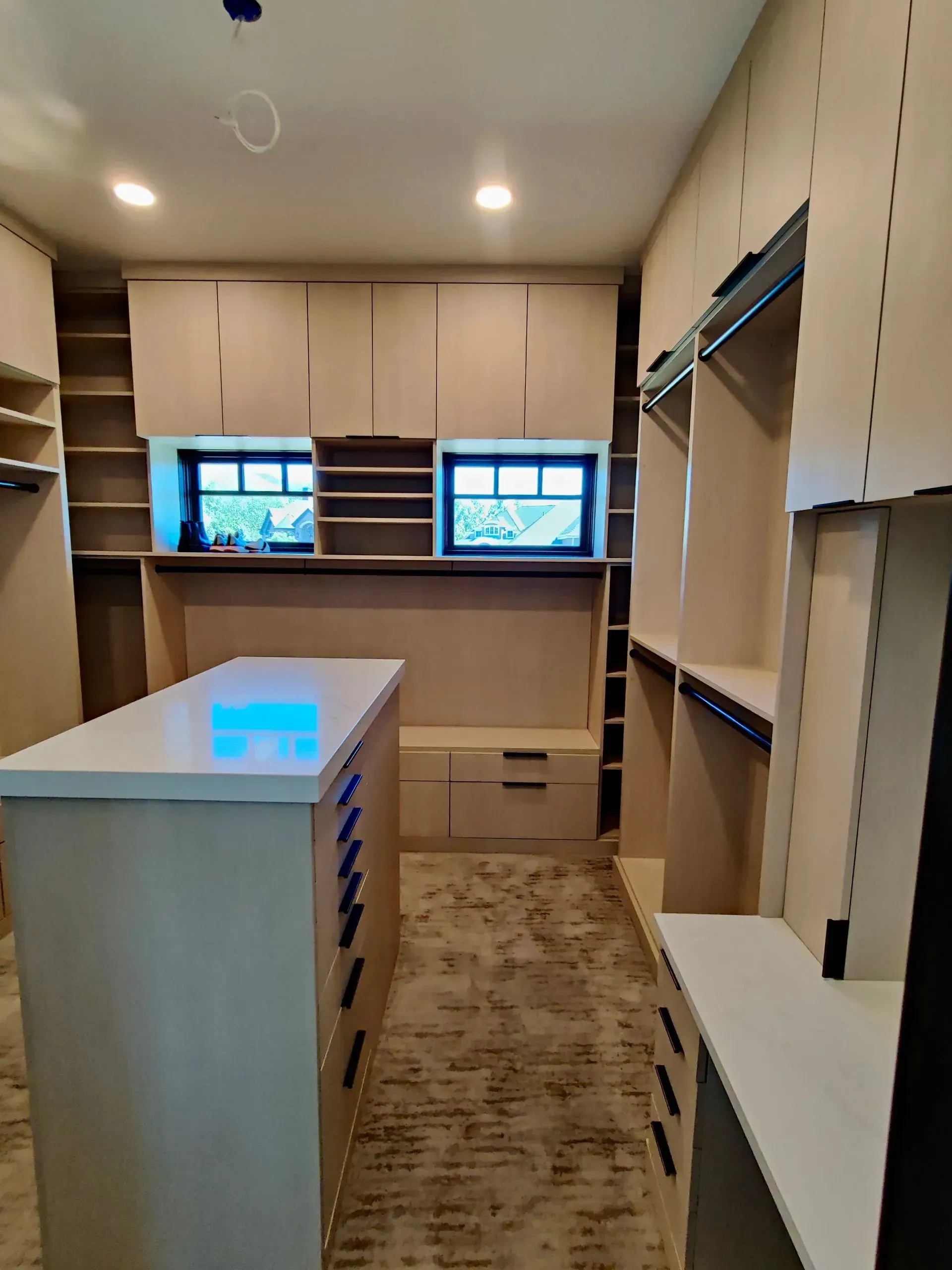 A modern walk-in closet featuring light wood cabinetry, white countertops, built-in storage, and two small windows.