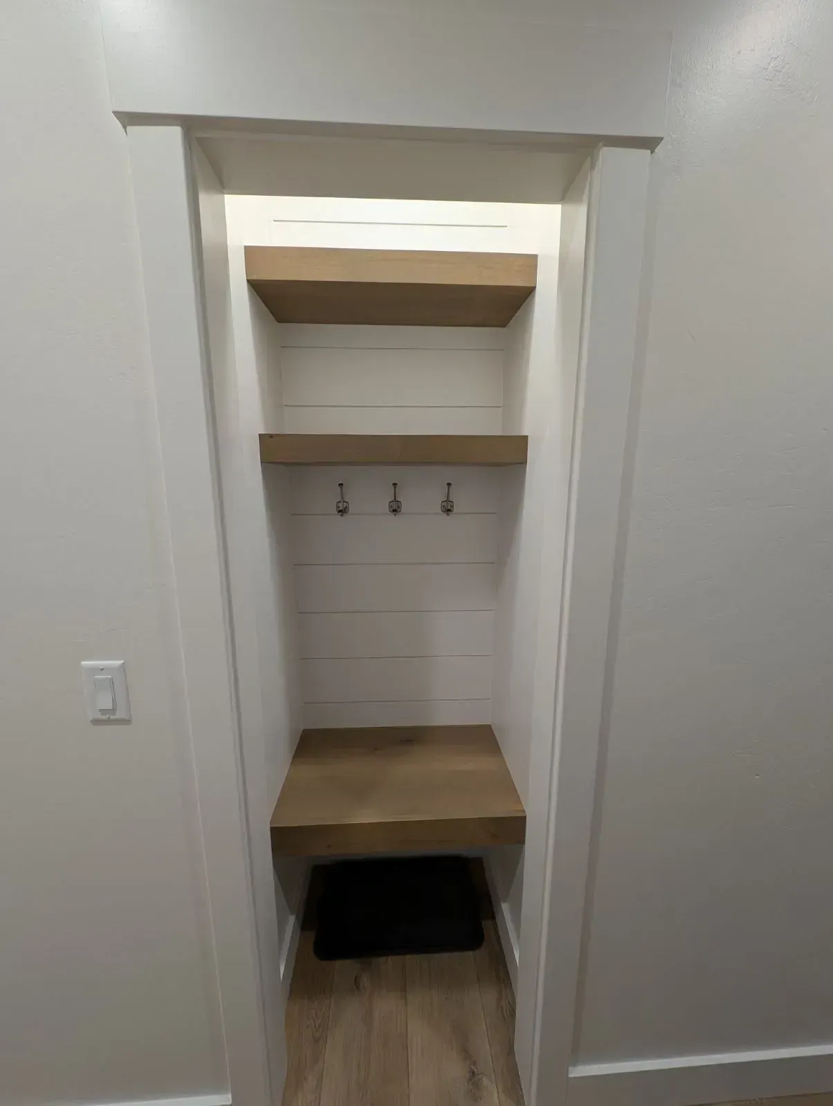 A built-in wall niche with light wood shelves, a bench, and coat hooks against a white shiplap-style backdrop.