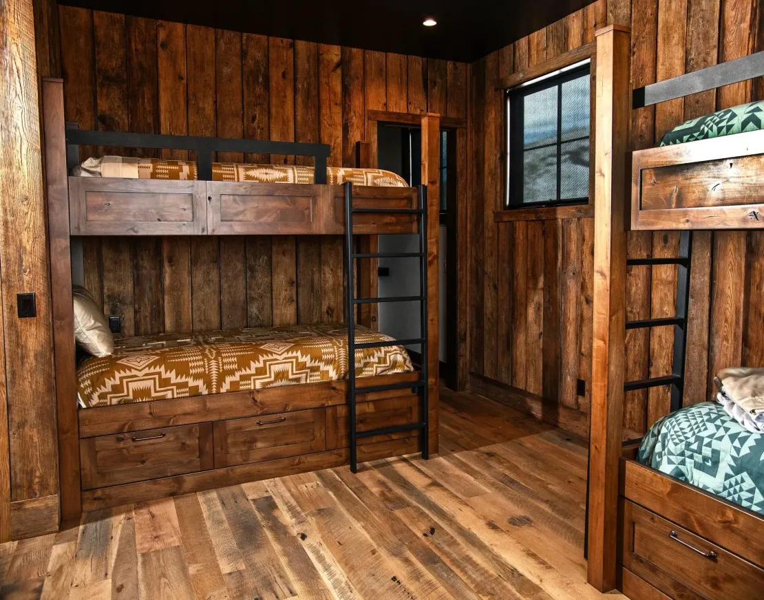 A rustic bedroom featuring two wooden bunk beds with patterned blankets against paneled wood walls and a dark ceiling.