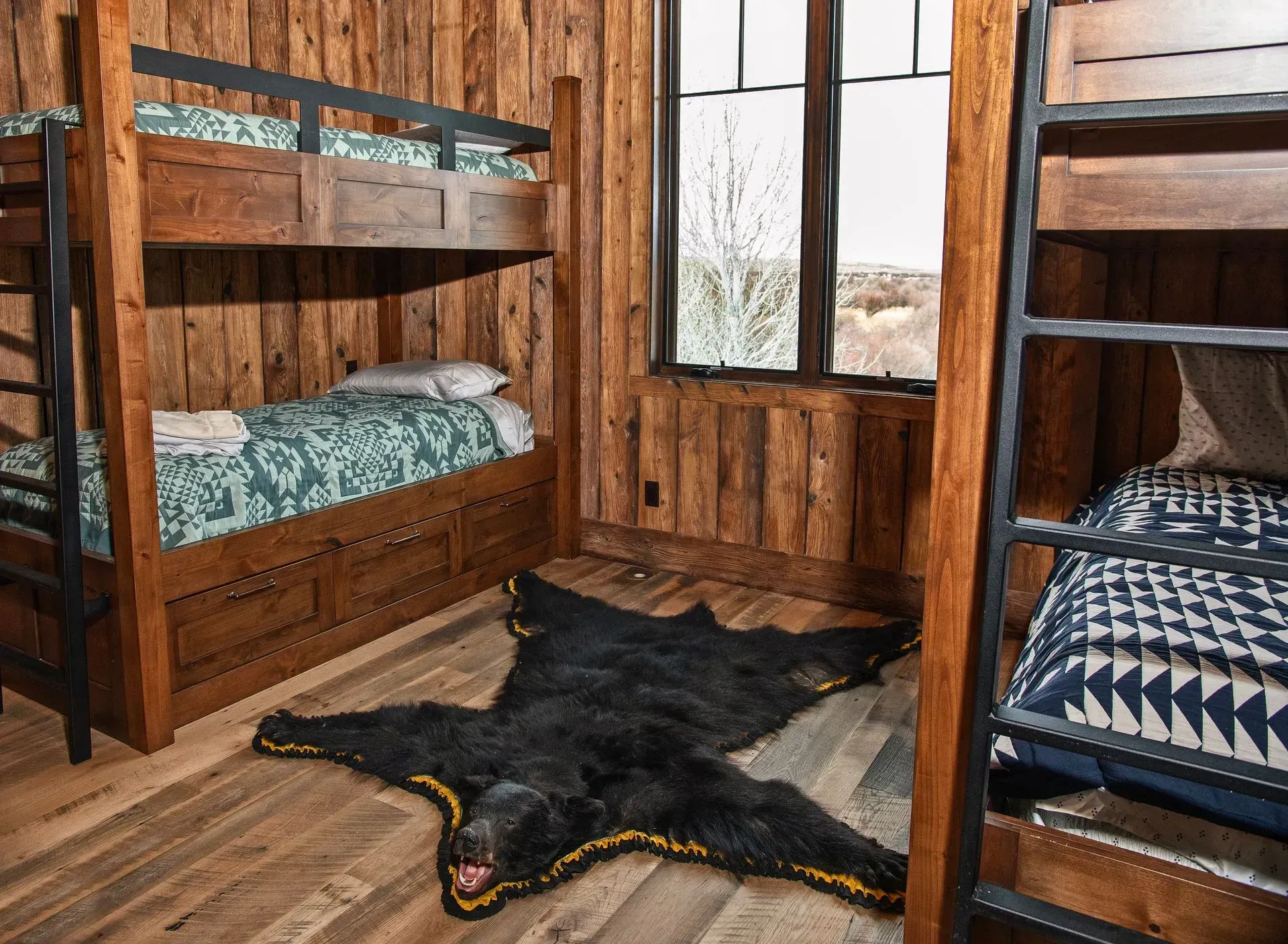 Rustic bedroom with two sets of bunk beds and a bear-skin rug on a wooden floor, next to a large window.