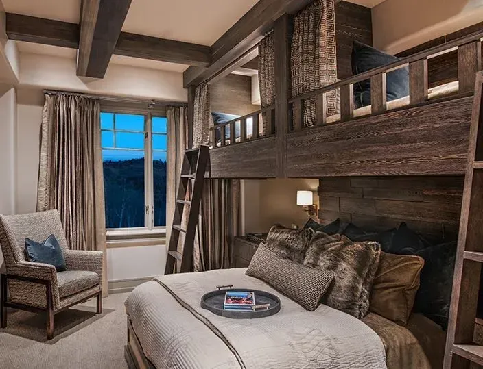 A rustic bedroom with a queen bed, wooden bunk beds above, a cozy armchair, and a large window at dusk.