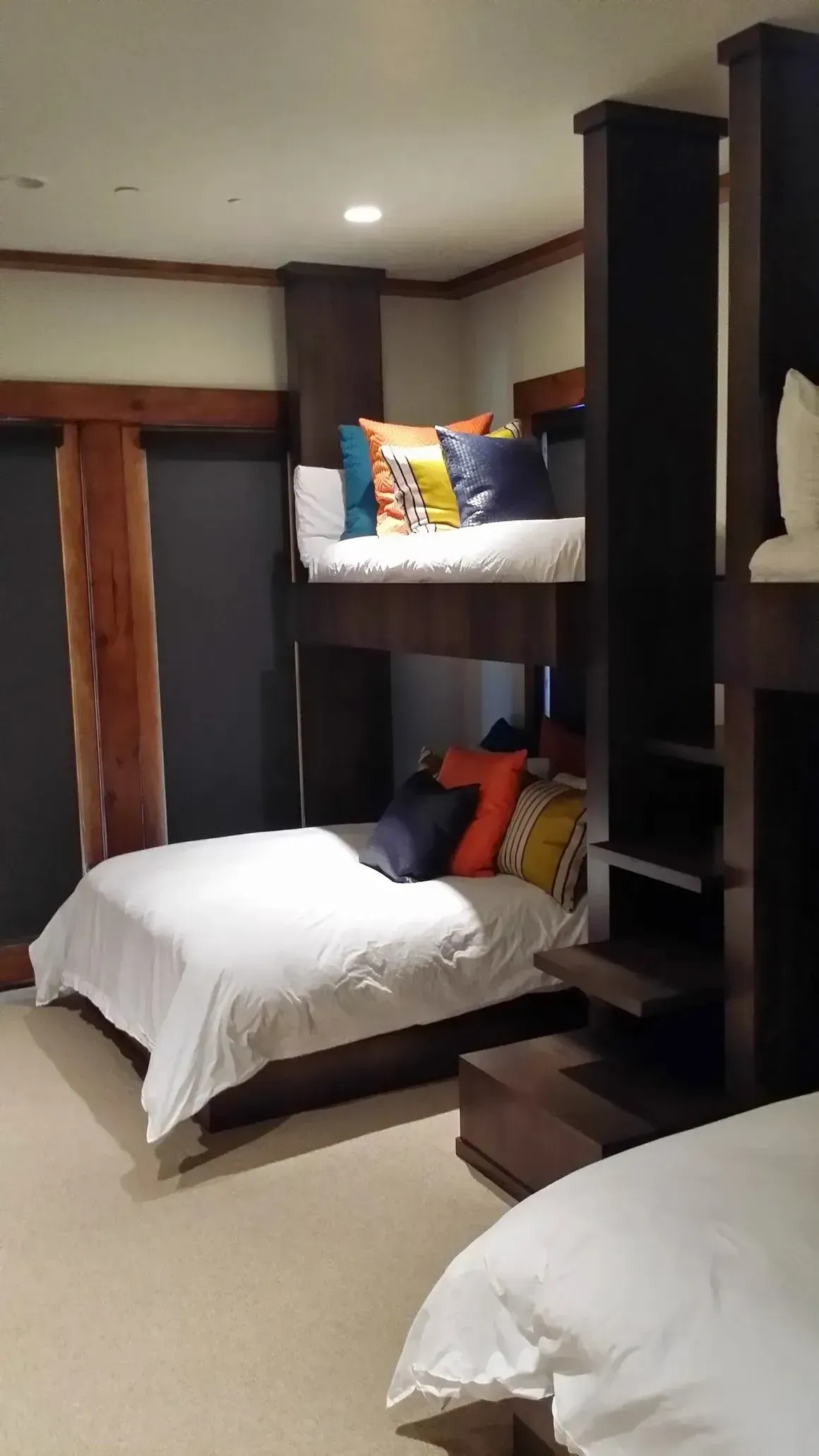 A room with built-in dark wood bunk beds, featuring white bedding, colorful accent pillows, and neutral carpet flooring.