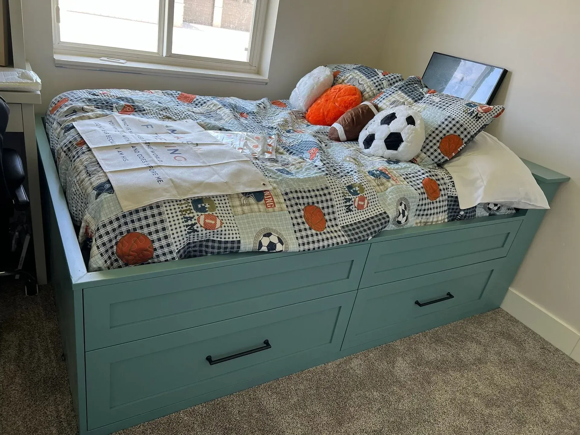 A light blue storage bed with sports-themed bedding and pillows, situated in a bedroom with carpeted flooring.