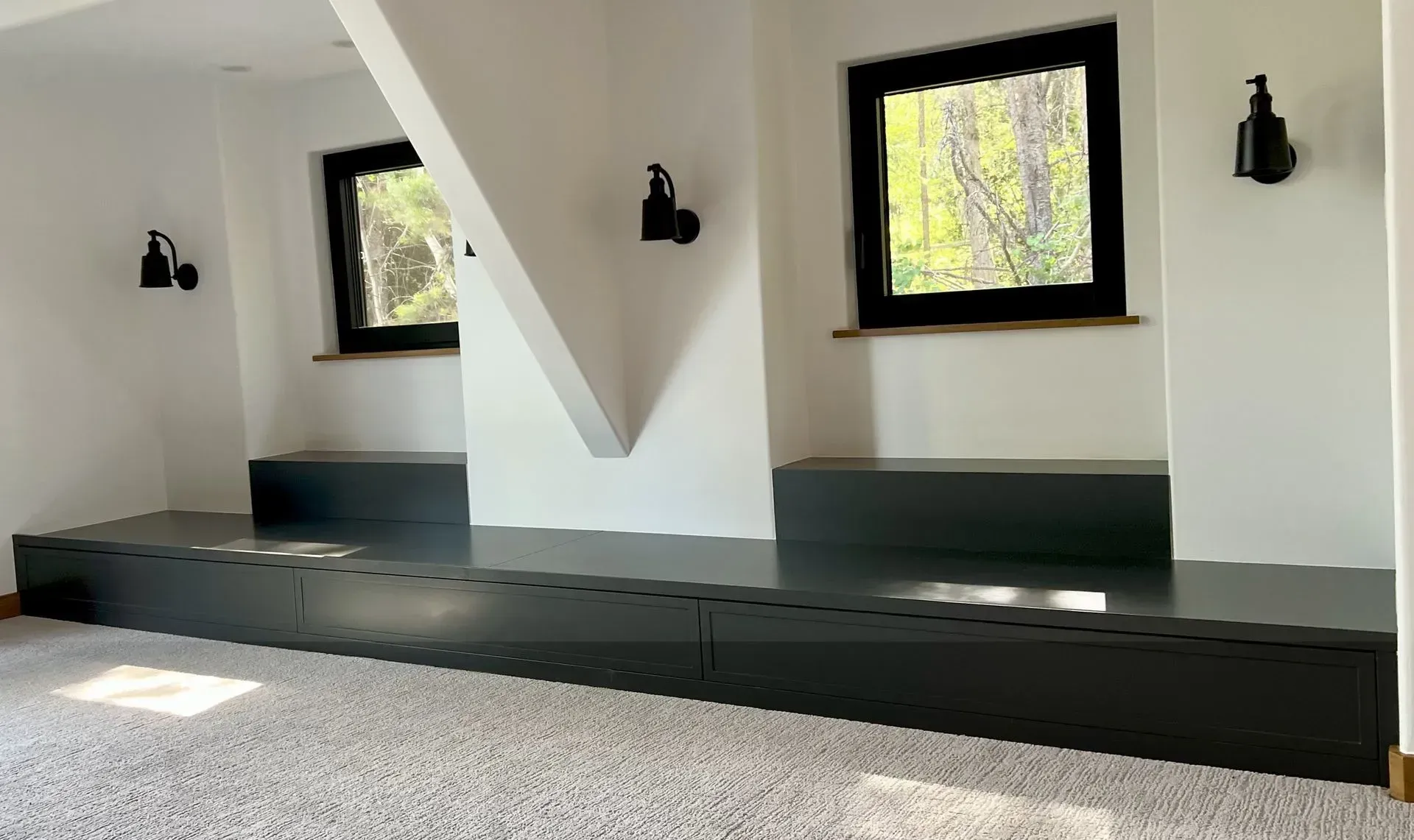 A white room with a low, black built-in bench spanning two recessed window nooks, with black wall sconces above each.