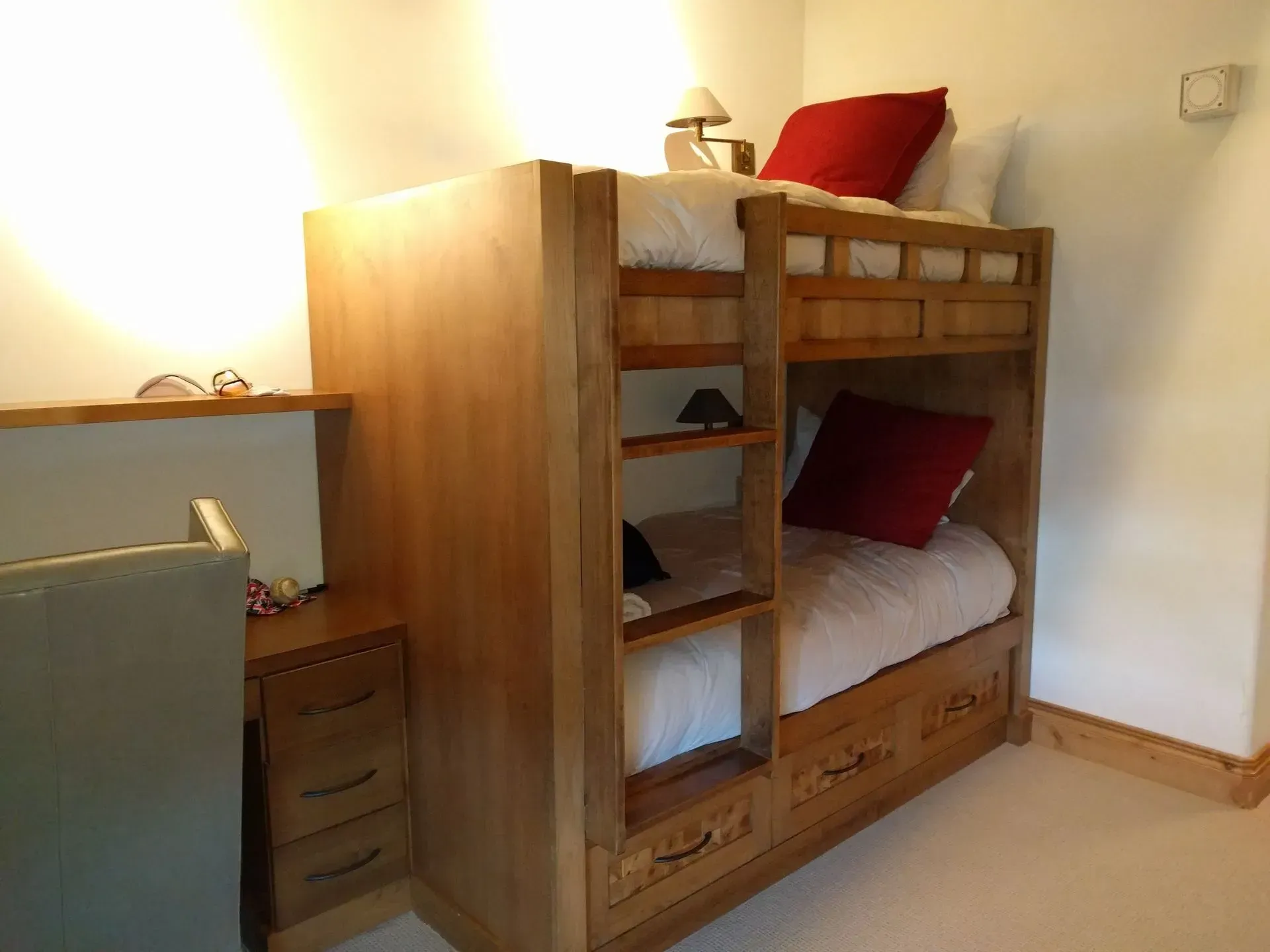 A wooden bunk bed with storage drawers, red pillows, and a small desk with drawers in a brightly lit room.