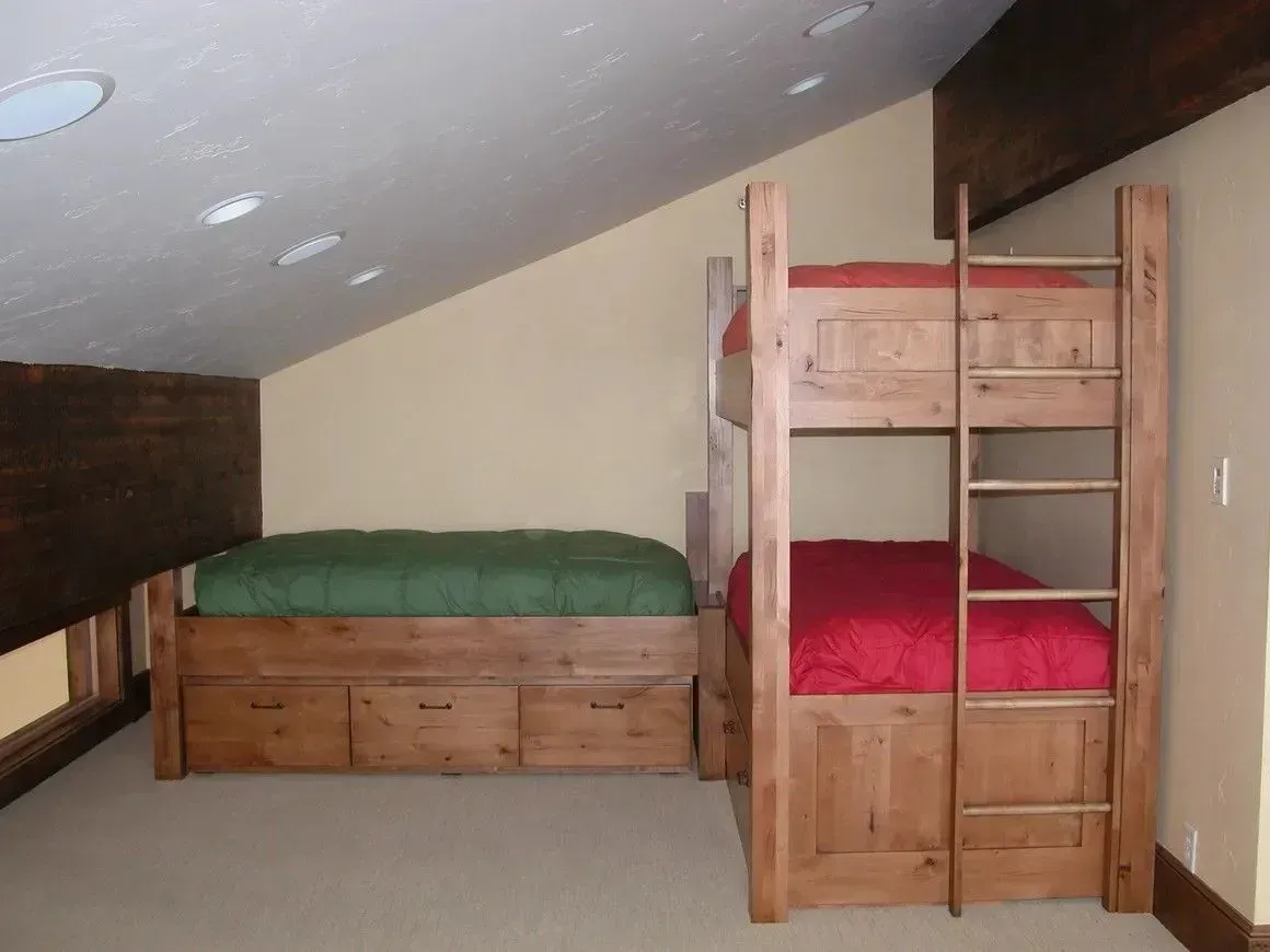 A room with a wooden bunk bed and a separate single bed with a green mattress on a sloped ceiling loft.