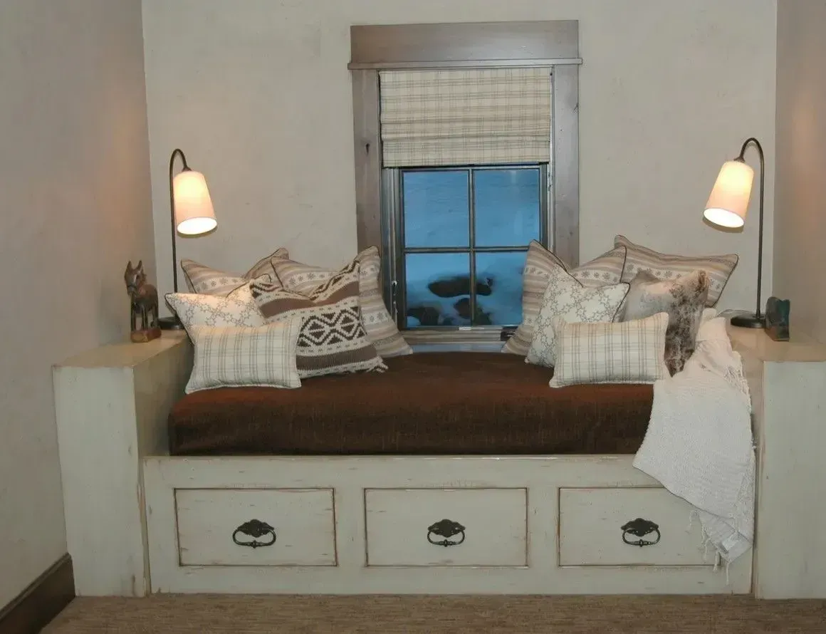 A built-in bench seat with storage drawers, brown cushion, and decorative pillows under a window with two reading lamps.