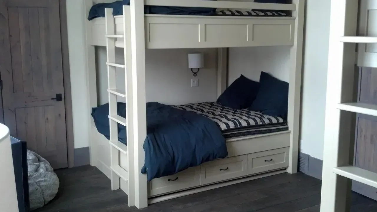 A cream-colored wooden bunk bed with navy bedding and storage drawers, positioned against a wall in a room with a door.