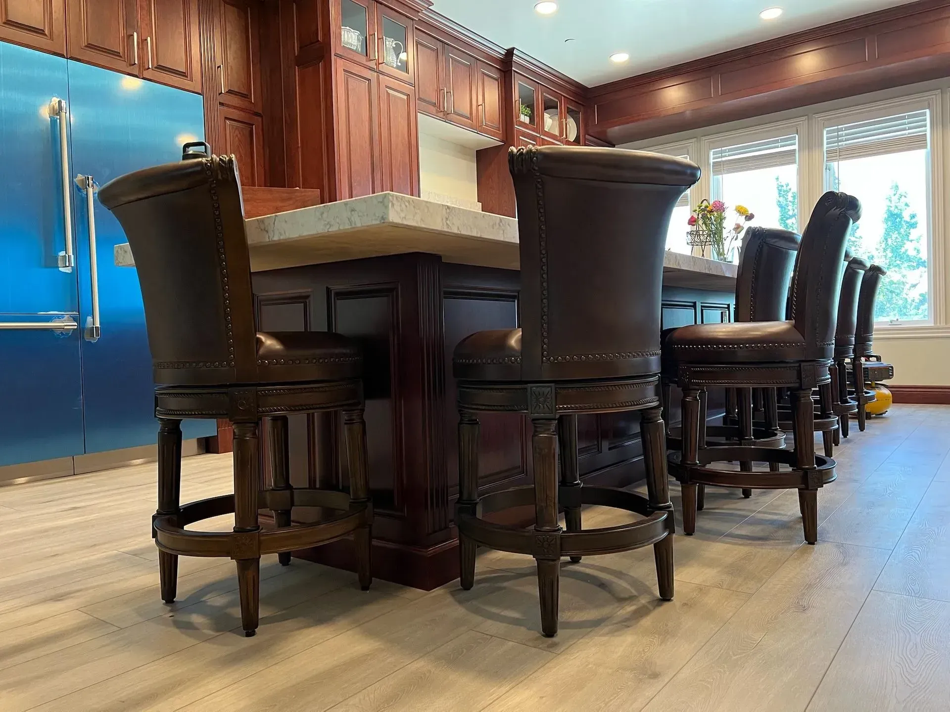 Dark brown bar stools line a wooden kitchen island with a stone countertop, situated in a kitchen with light wood floors.