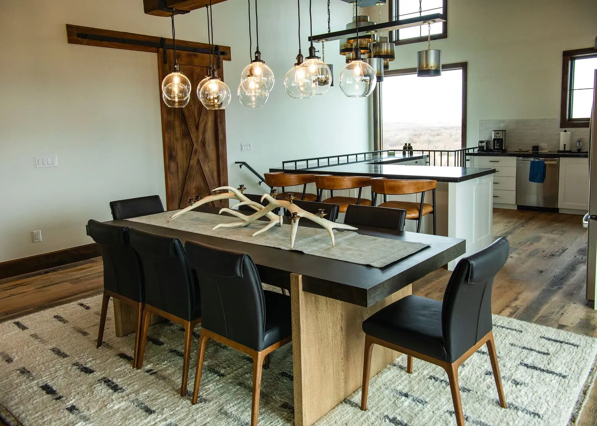 A dining area with a modern dark table, six black chairs on a patterned rug, pendant lights, and a kitchen background.