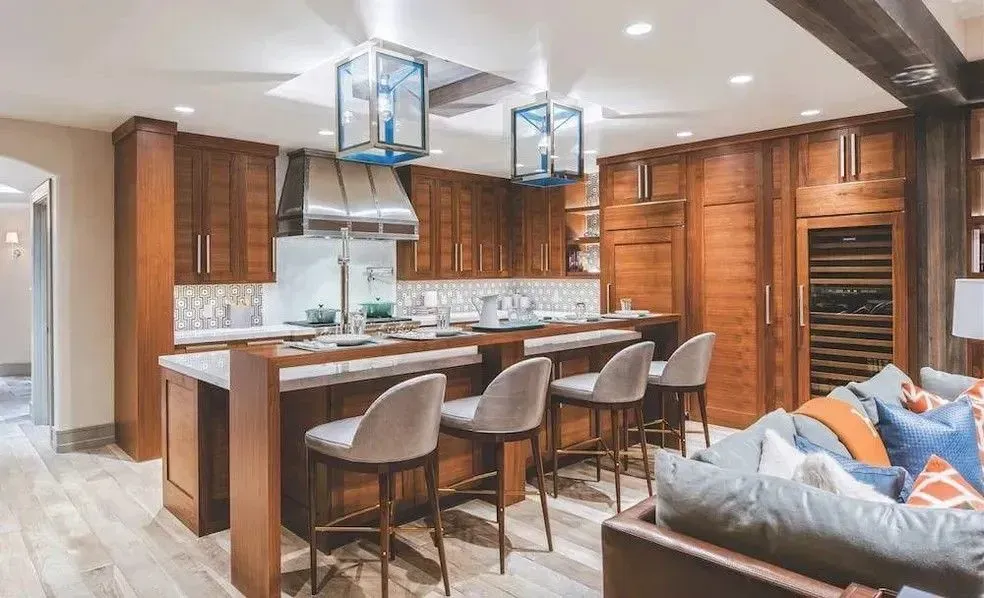 A modern kitchen featuring wooden cabinets, a long breakfast bar with barstools, and two large hanging lantern lights.