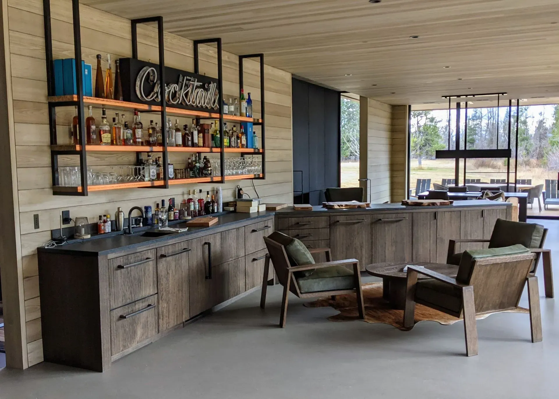 A modern home bar with wooden cabinets, open industrial shelving filled with bottles, and two wood-framed armchairs.