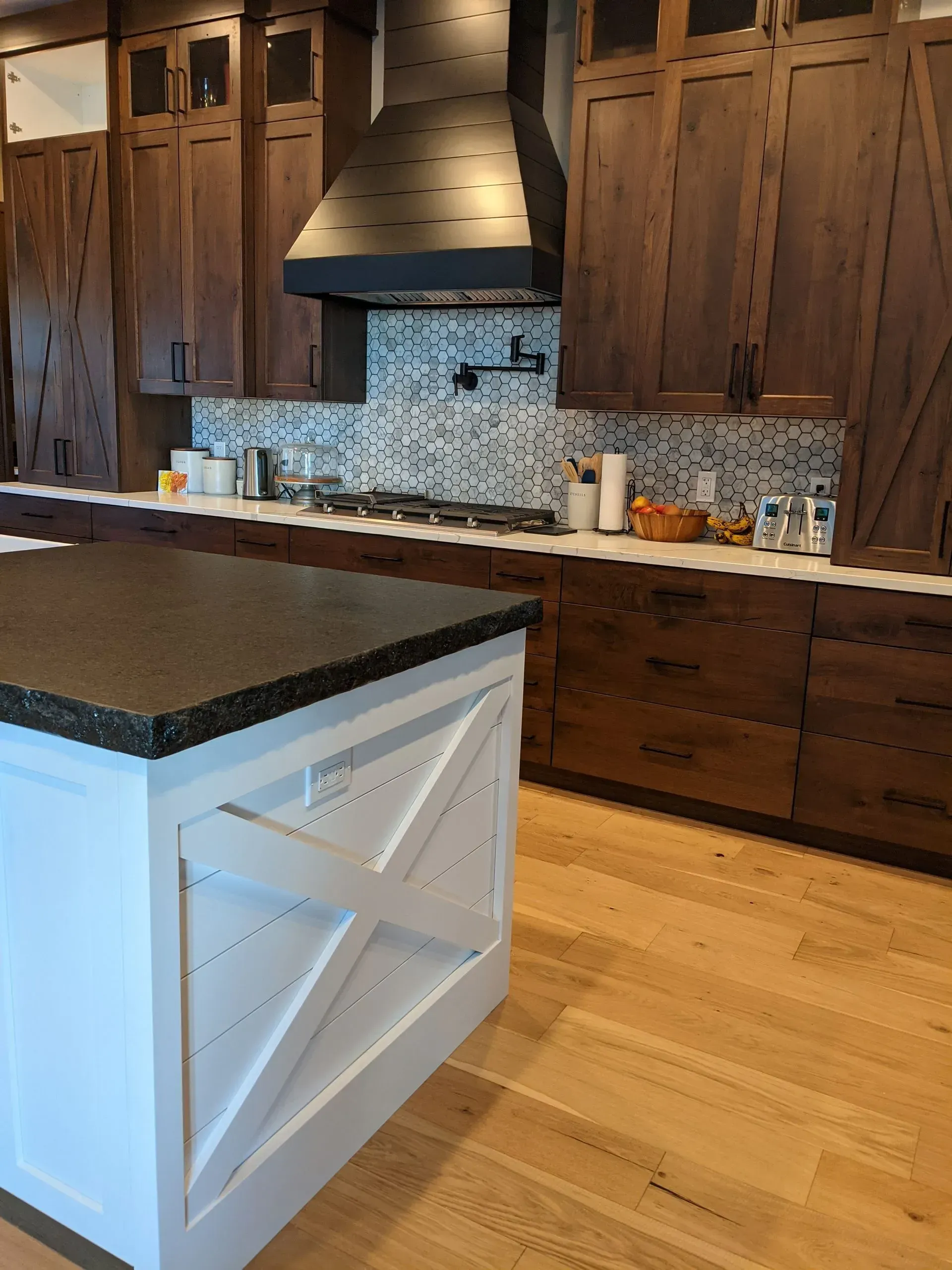 A kitchen with white farmhouse-style island cabinetry, dark wood cabinets, a metal vent hood, and light wood flooring.