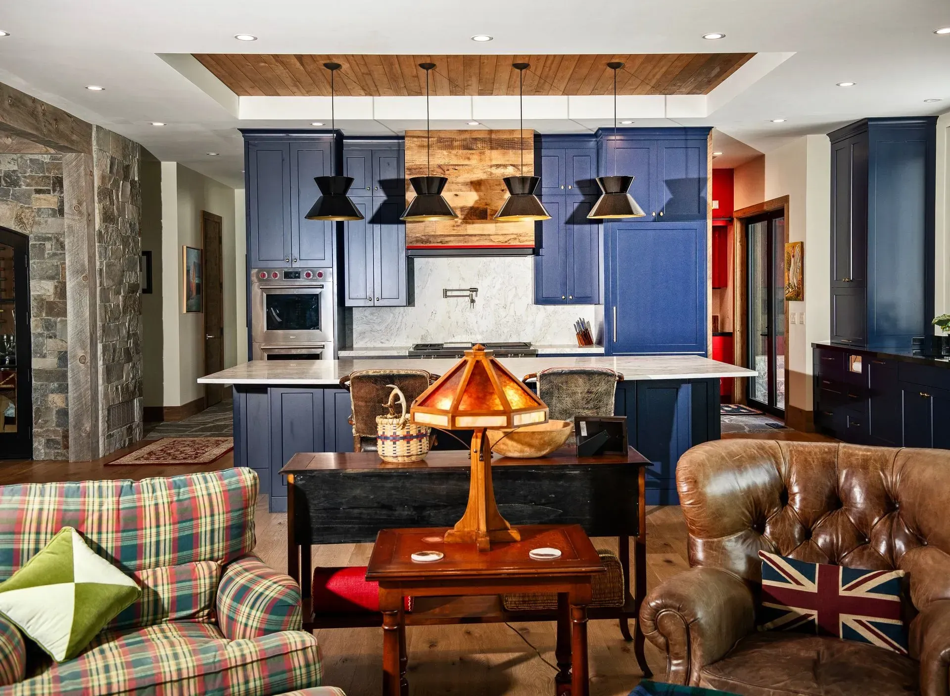 A rustic living room with a plaid chair and leather sofa, opening to a modern blue kitchen with a wooden ceiling feature.