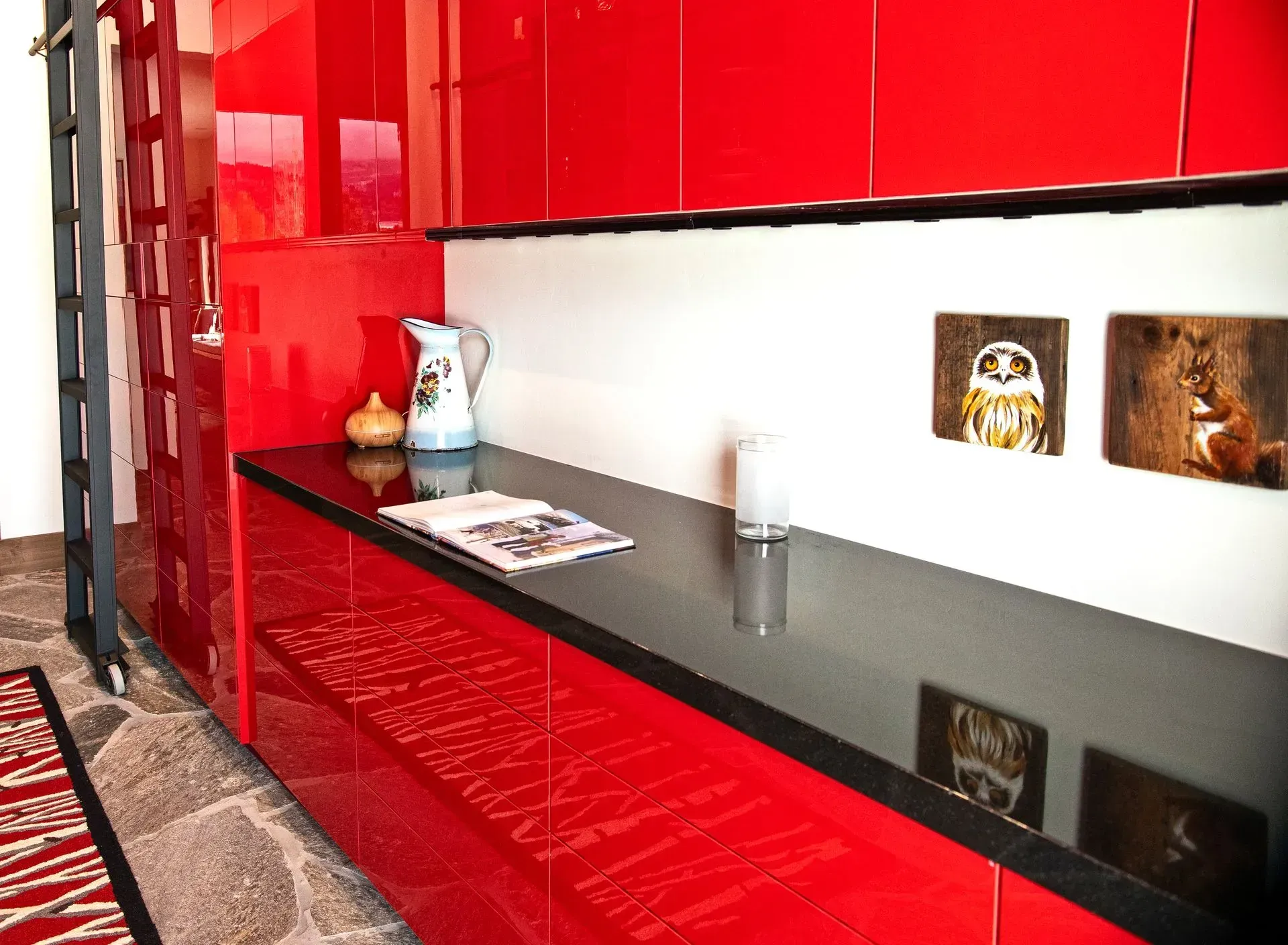 A black counter sits below bright red cabinets and two small animal photos on a white wall, with a pitcher and book.