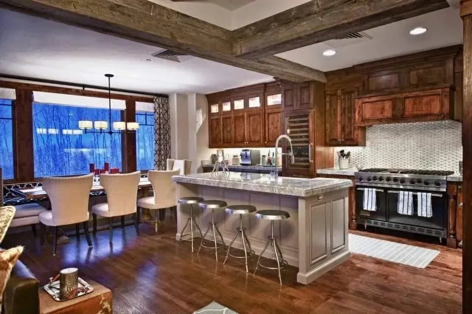 A kitchen with wood cabinets, a light-colored island with three metal bar stools, and an adjacent dining area with a window.