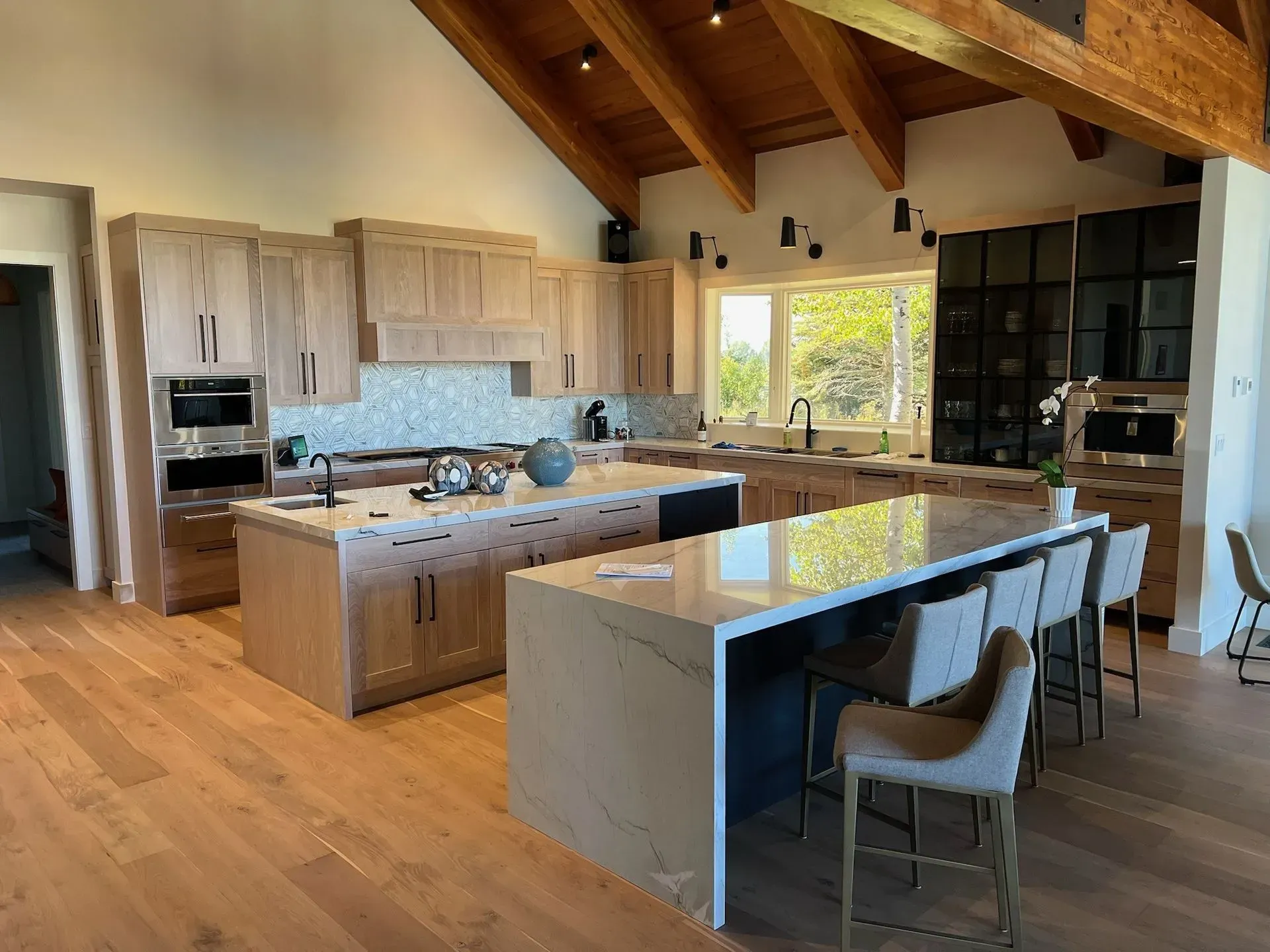 A modern kitchen with light wood cabinetry, two large islands with stone countertops, and high-vaulted ceilings.