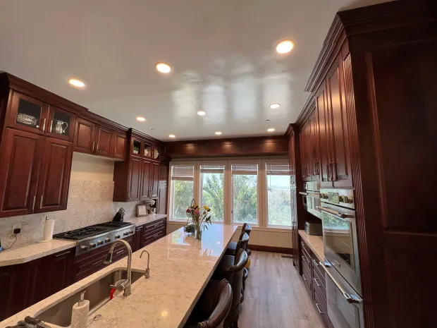 A spacious, modern kitchen with dark wood cabinetry, a long marble-topped island, a stove, and large windows.