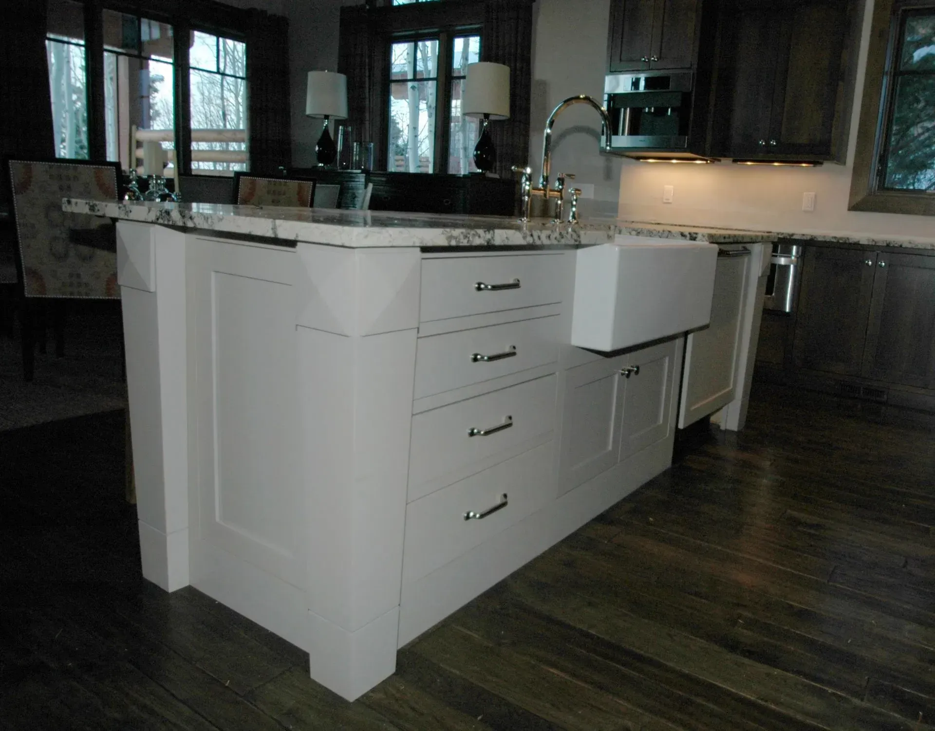 A white kitchen island featuring a farmhouse sink, drawers, and a marble countertop set in a dark wood kitchen.