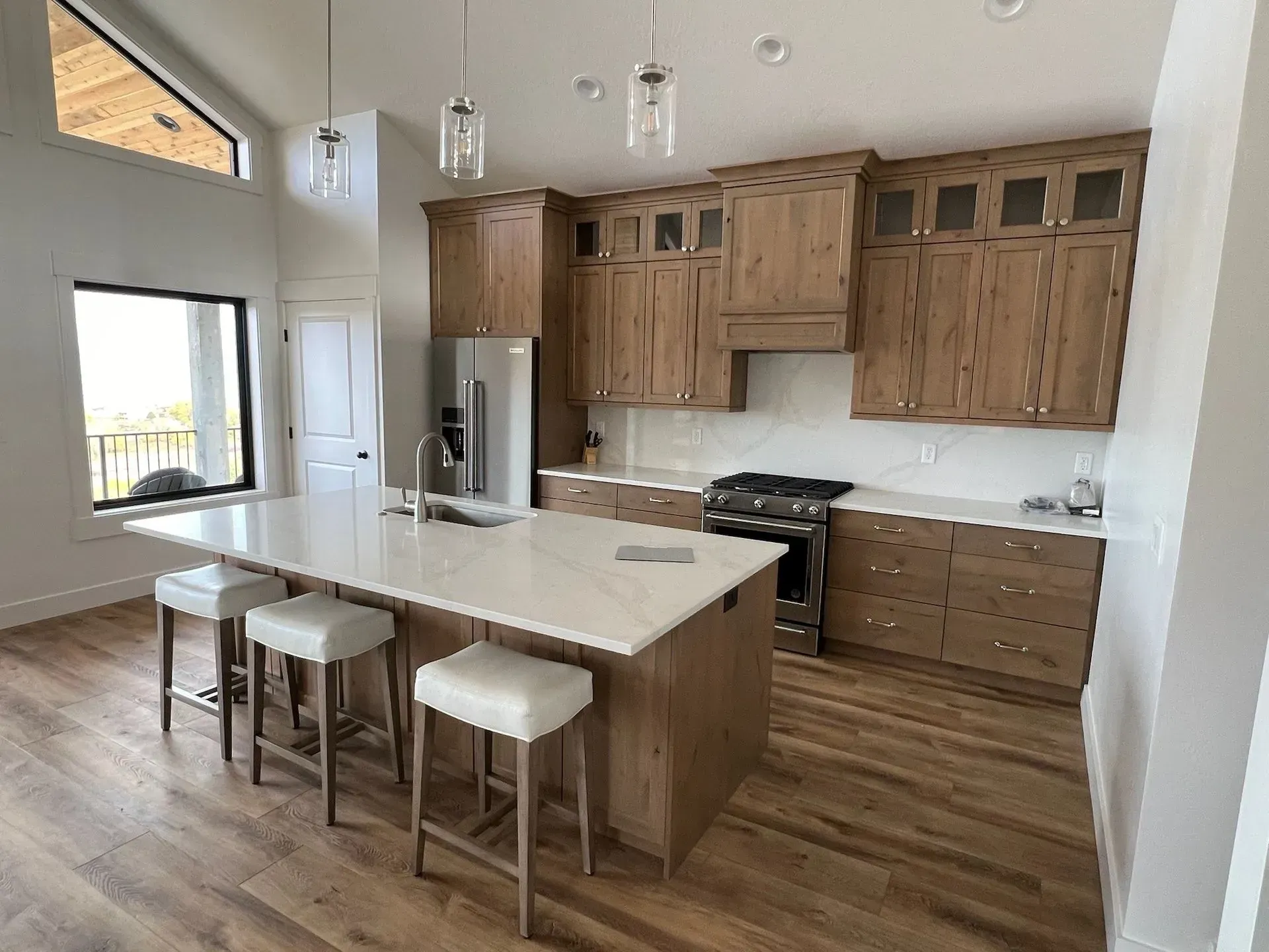 Modern kitchen with wood cabinets, quartz island with three white stools, and hardwood floors in a sunlit open space.