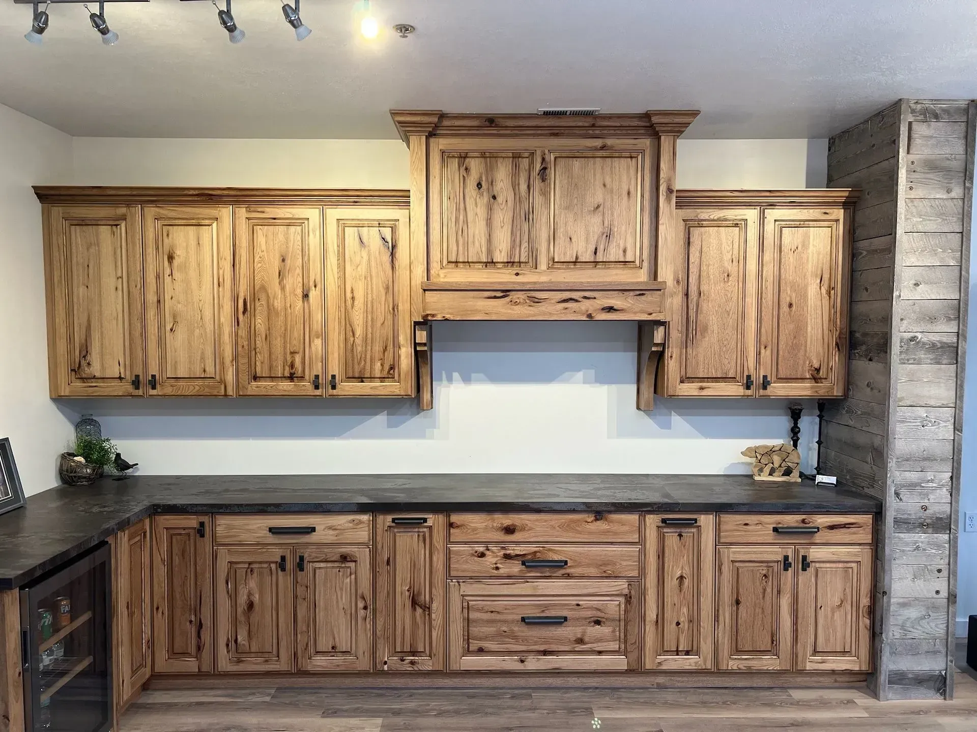 A kitchen with rustic knotty alder wood cabinets, dark countertops, and a tall wooden range hood against a light wall.