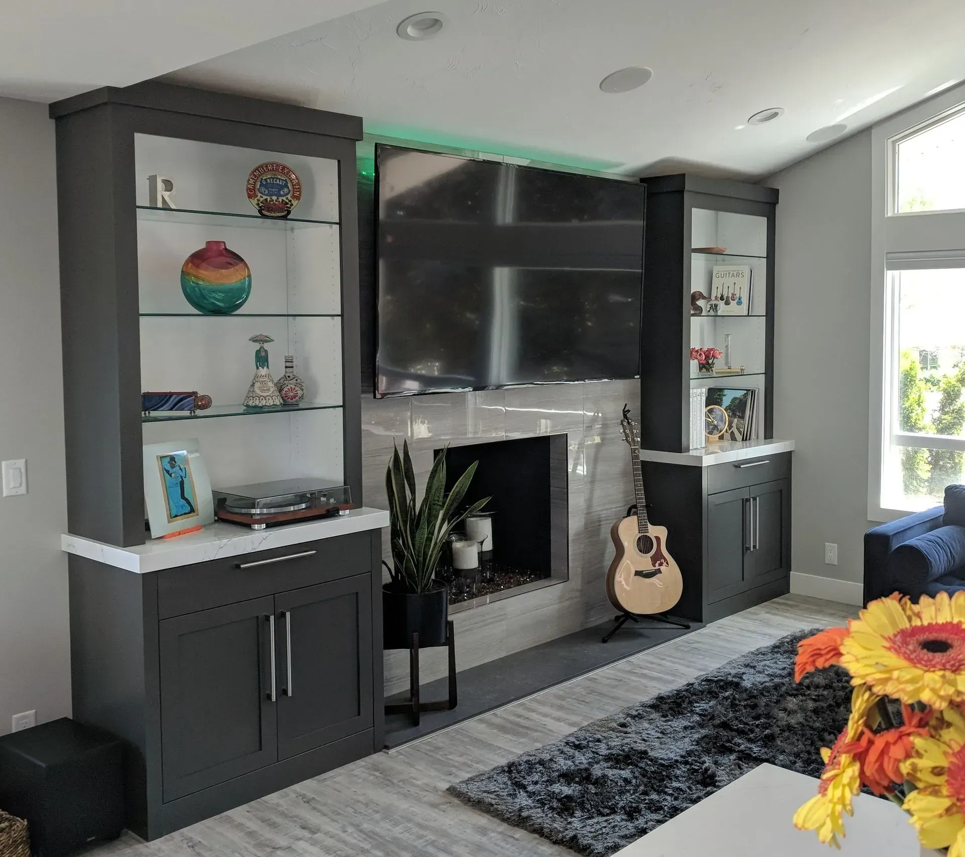 A large flat-screen TV above a fireplace, flanked by dark gray storage cabinets with glass shelving and home decor.